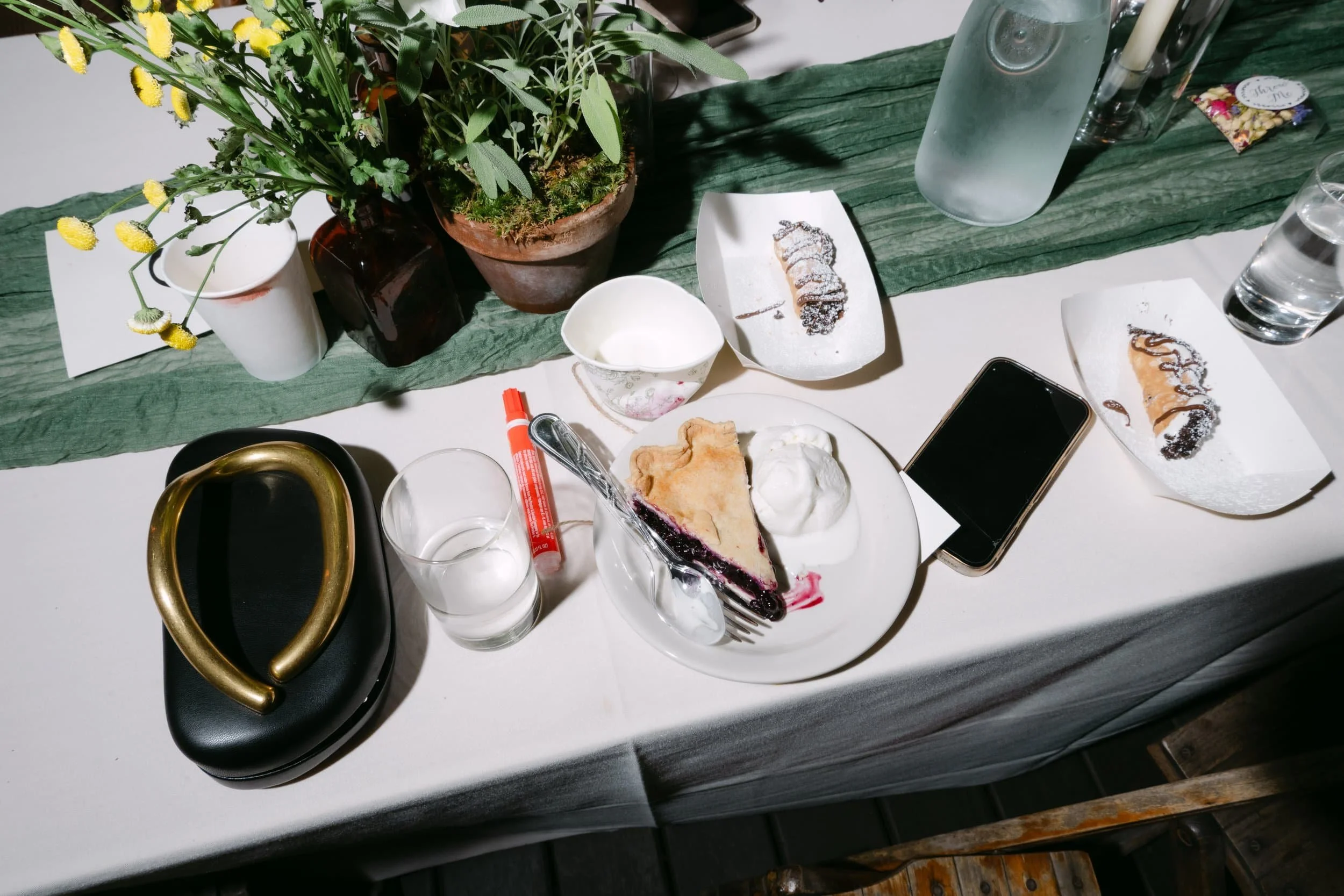 A table with a white tablecloth, green runner, and various items including a potted plant, a black purse with a gold handle, a clear glass of water, a slice of blueberry pie with whipped cream, empty bowls, a smartphone, a bottle of water, and plates with leftover cake and chocolate drizzle.