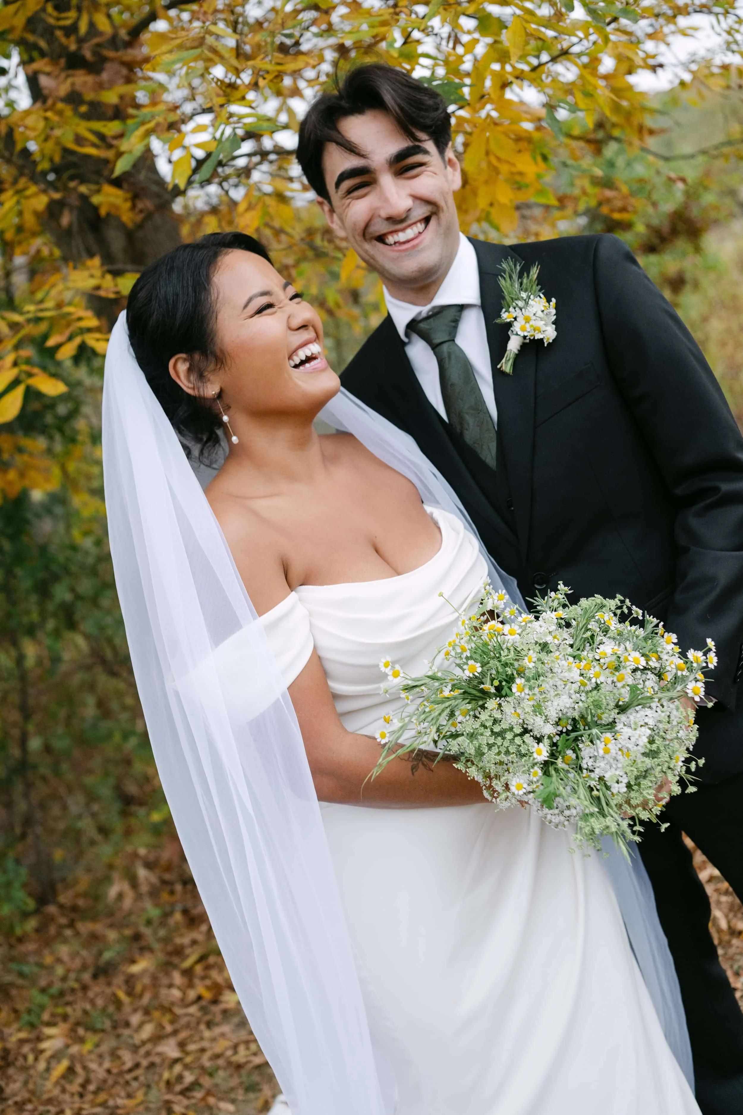 A joyful bride and groom laughing outdoors with autumn foliage in the background. The bride is holding a bouquet of wildflowers, wearing an off-shoulder white wedding dress and a veil. The groom is in a black tuxedo with a boutonniere, leaning towards the bride.