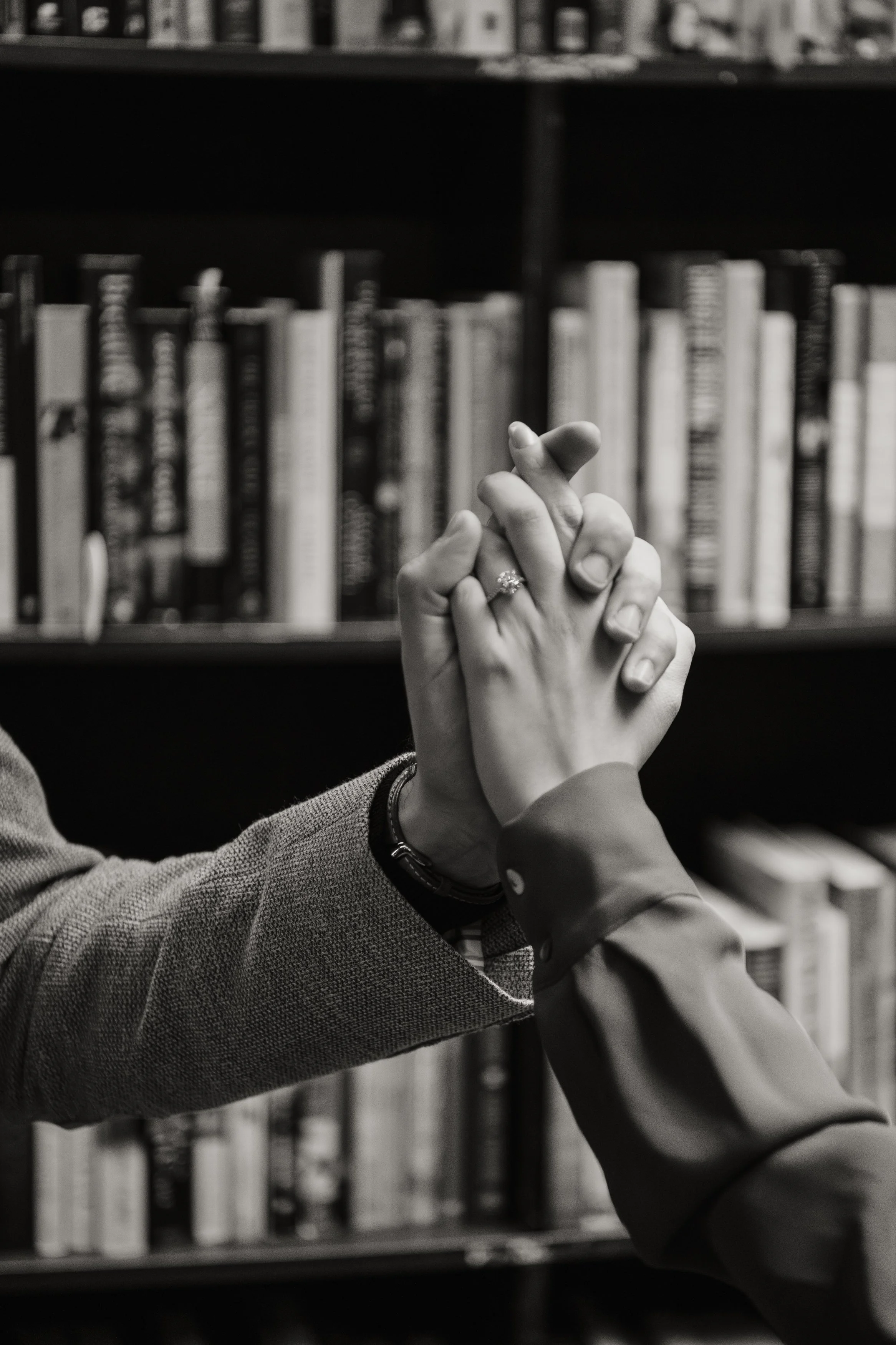 Two people clasp hands in front of a bookshelf, one wearing a ring, in a black and white photo.