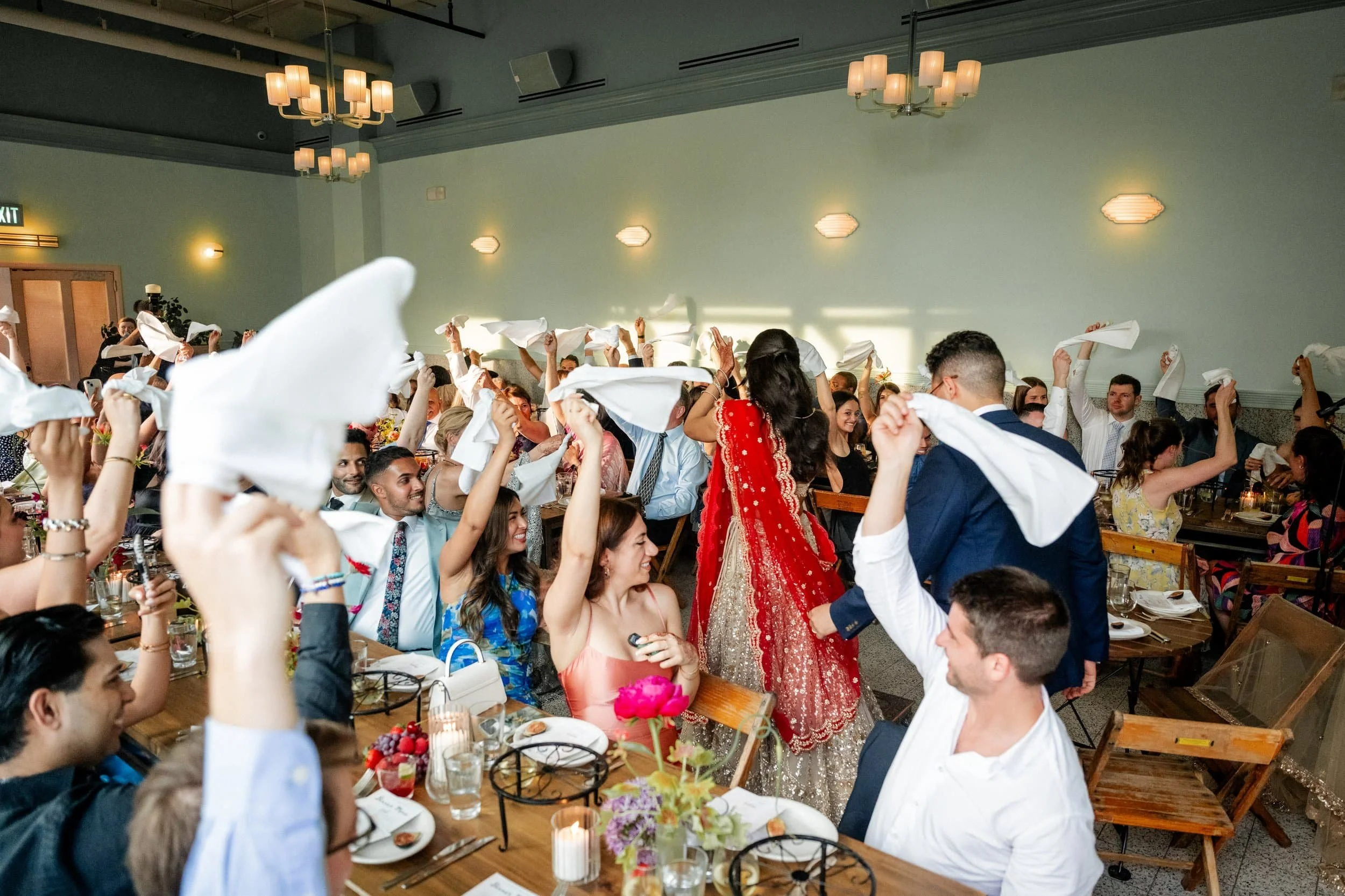 People celebrating at a wedding reception, including a woman in a red dress in the center, as guests wave white napkins in the air.