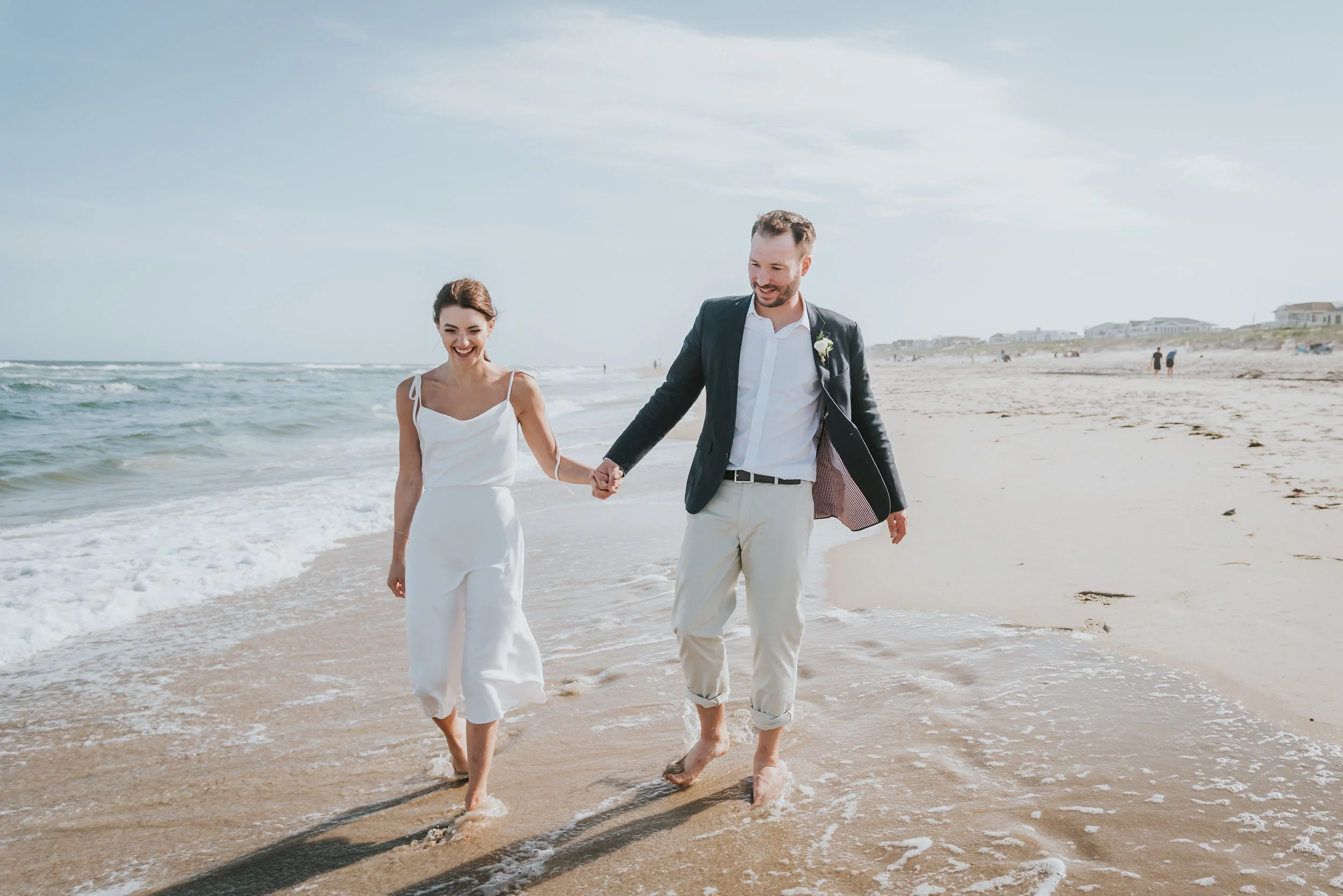 A couple dressed in wedding attire walking hand in hand along the beach, with waves washing over their feet and smiling at each other.