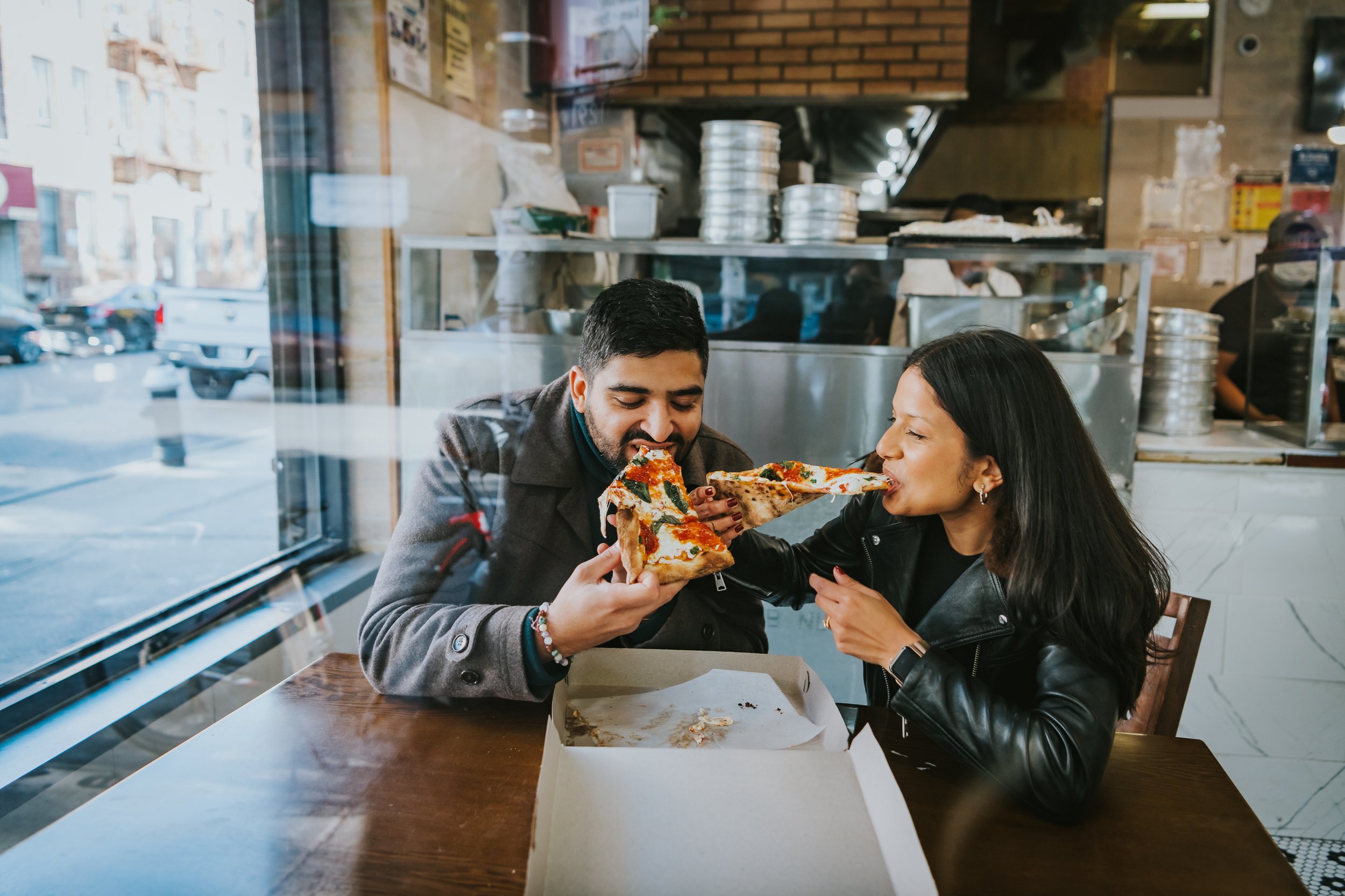 Two people sharing a slice of pizza inside a pizzeria, seen through a large window with street view.