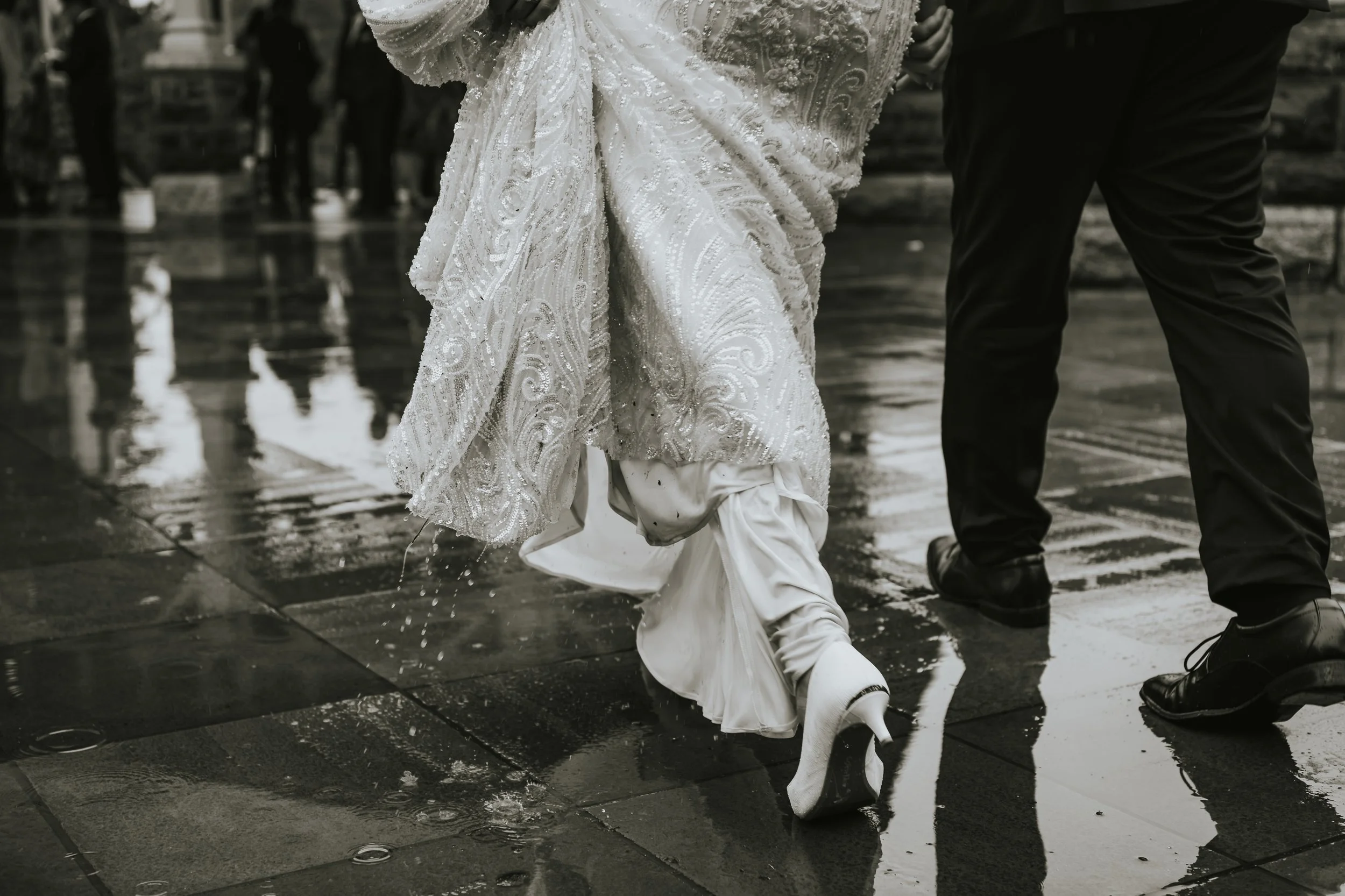 Bride in a wedding dress and high heels walking on wet pavement, with a groom in dress shoes, in the rain.