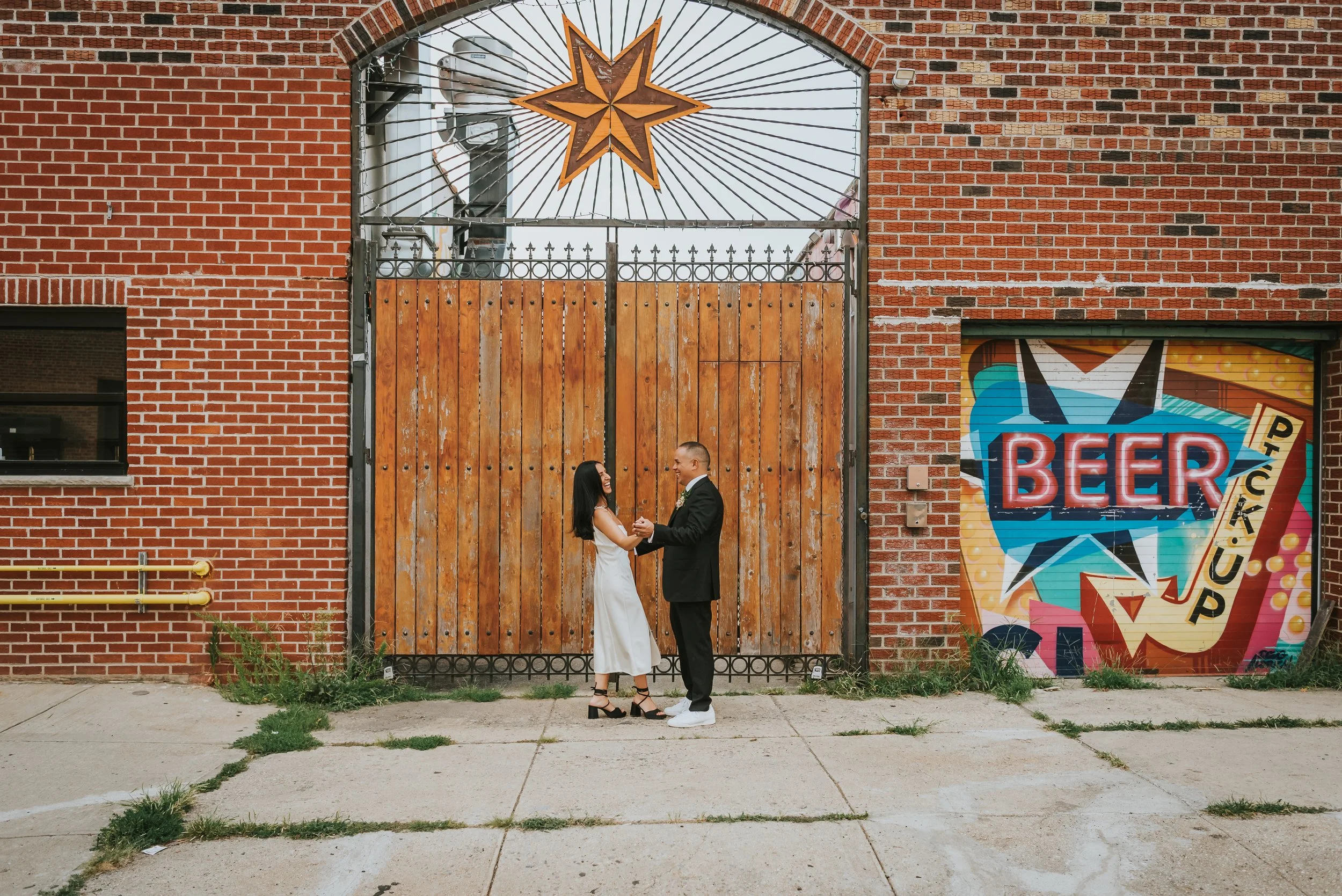 A couple stands holding hands and facing each other on a sidewalk in front of a brick building with a large wooden gate. The woman wears a white dress and black heels, and the man wears a black suit and white shoes. To the right, a colorful mural fea