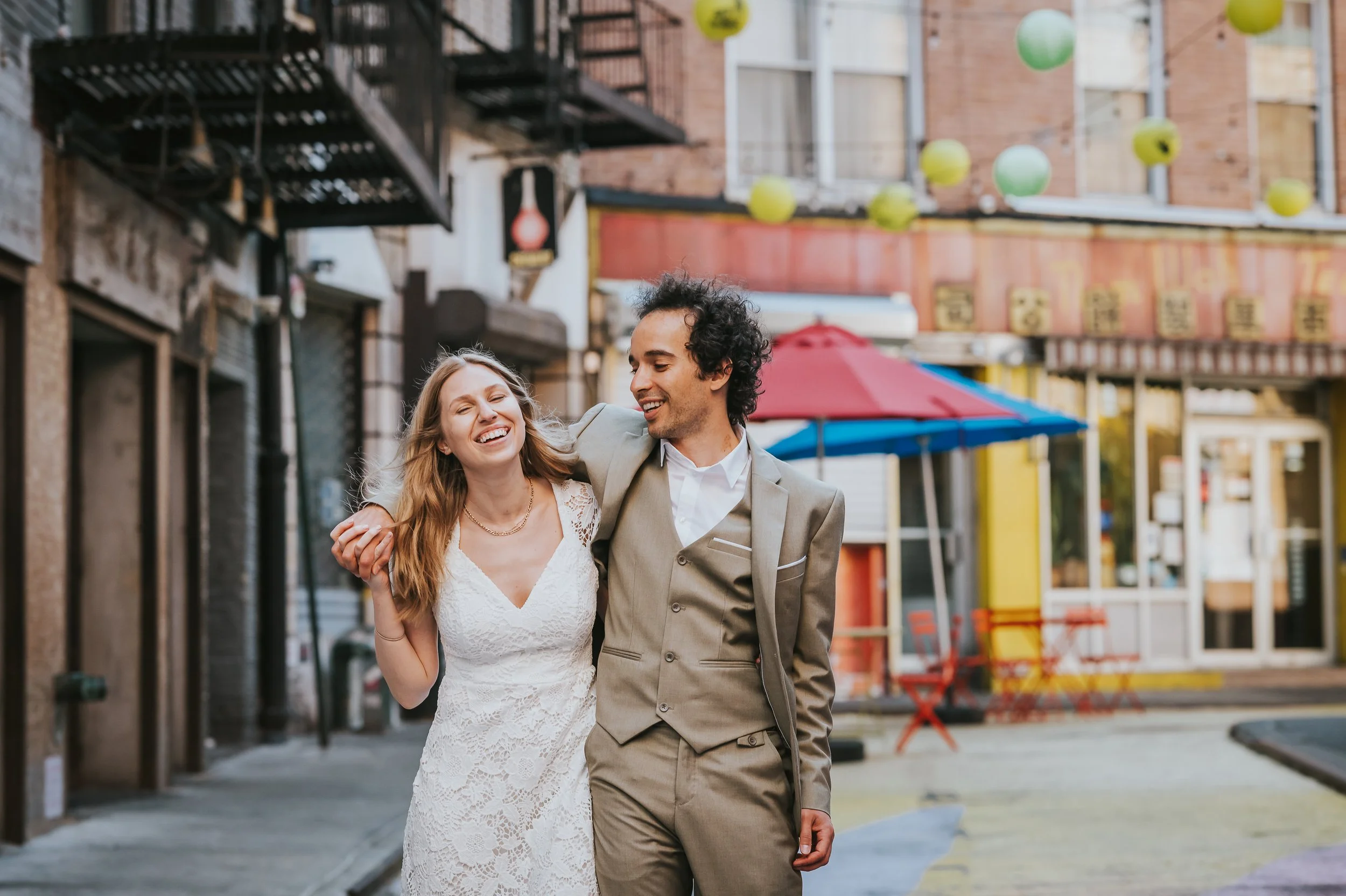 A smiling woman in a white lace dress and a man in a beige suit walk arm in arm on a lively city street decorated with colorful lanterns and umbrellas.