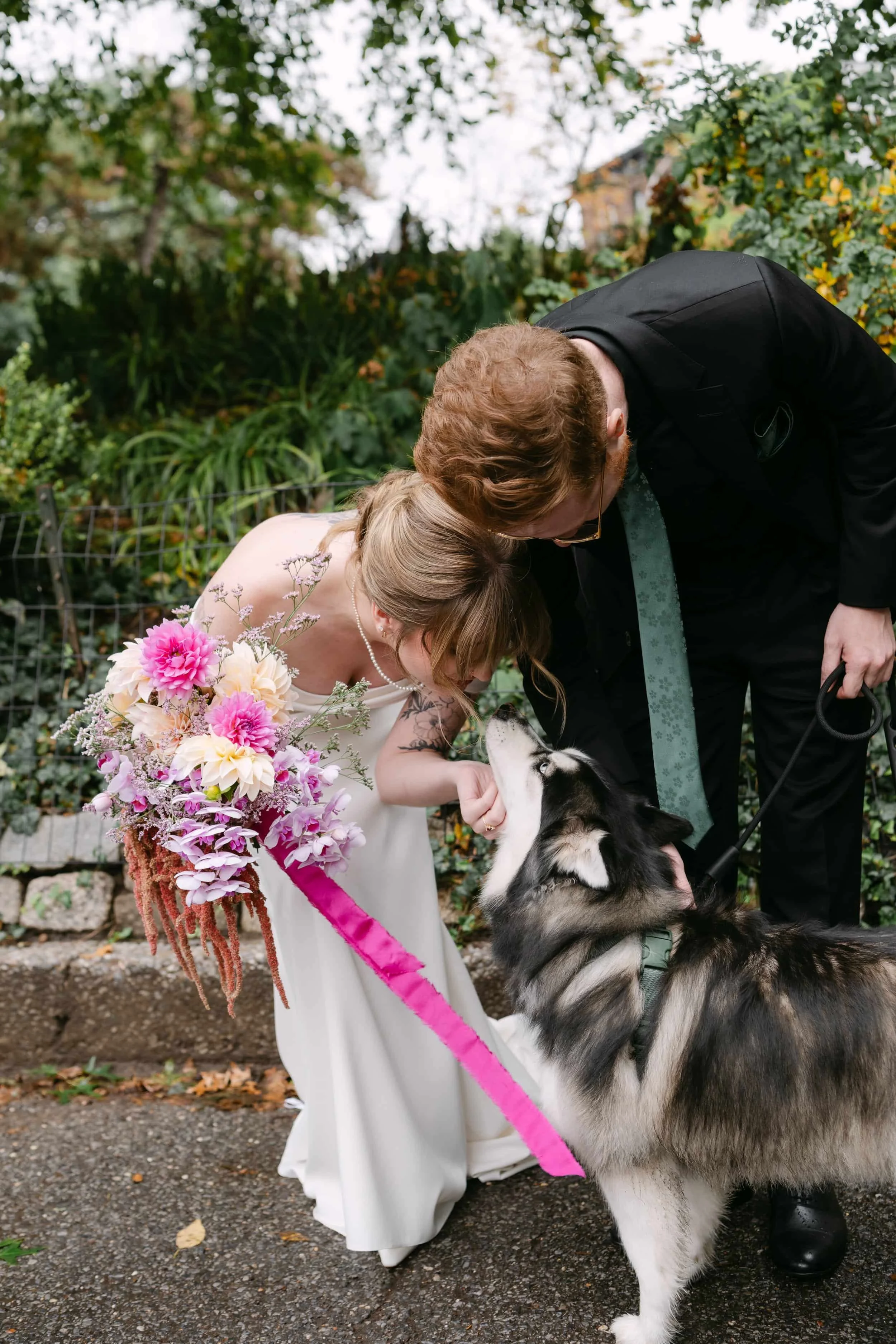 Bride in white dress holding a bouquet leaning towards a husky dog, with a man in black suit and tie kissing the bride on the head outdoors.