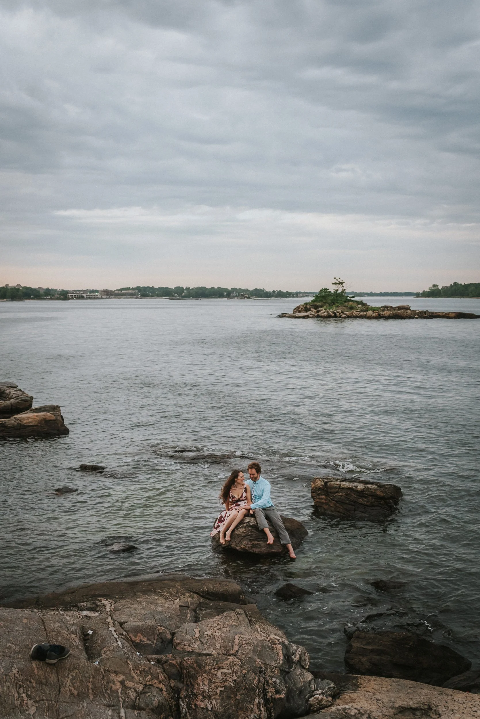 A couple sitting on a large rock in a lake under a cloudy sky, with smaller rocks around them and a small island in the distance.