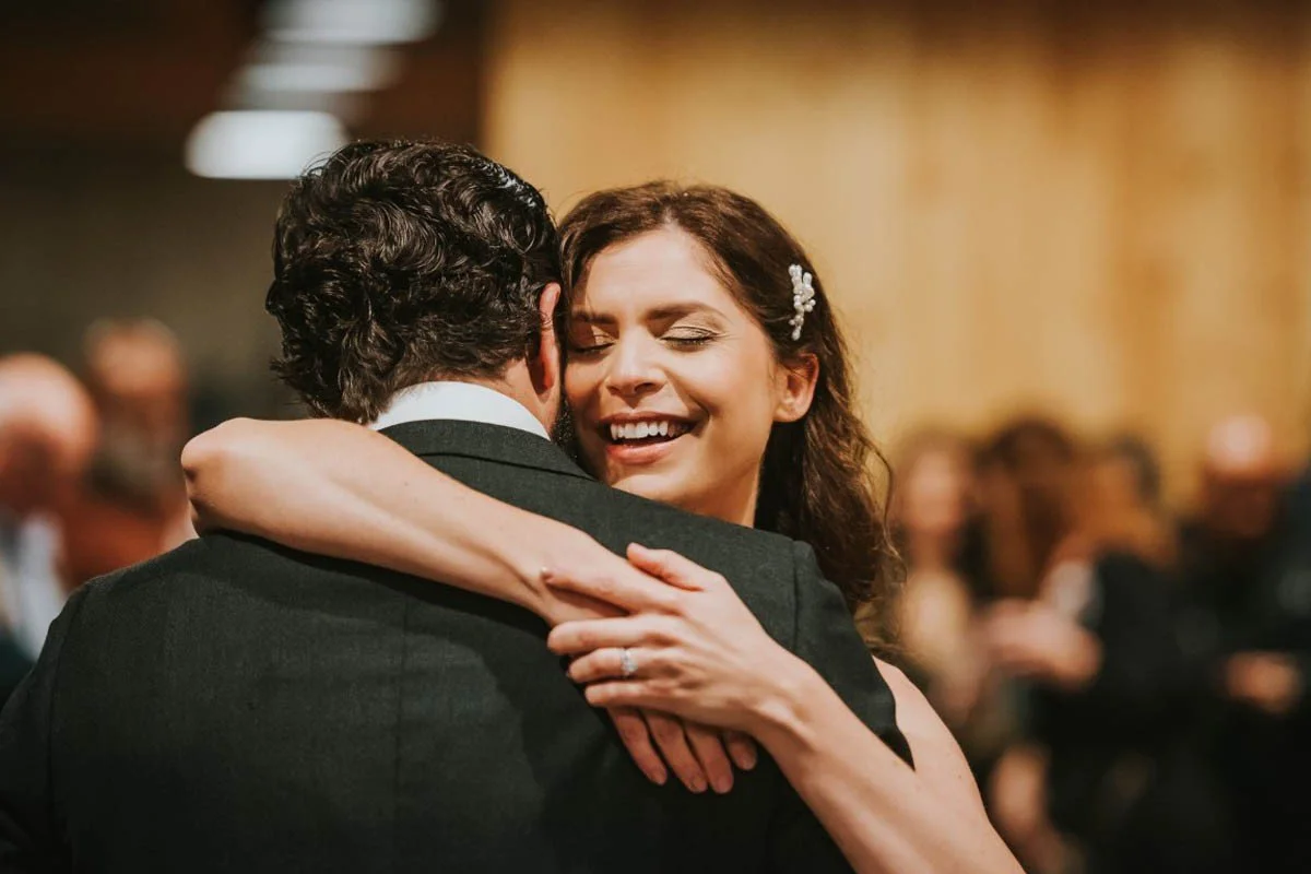 A woman in a black dress with a pearl hair accessory smiling and hugging a man in a tuxedo at a formal event.