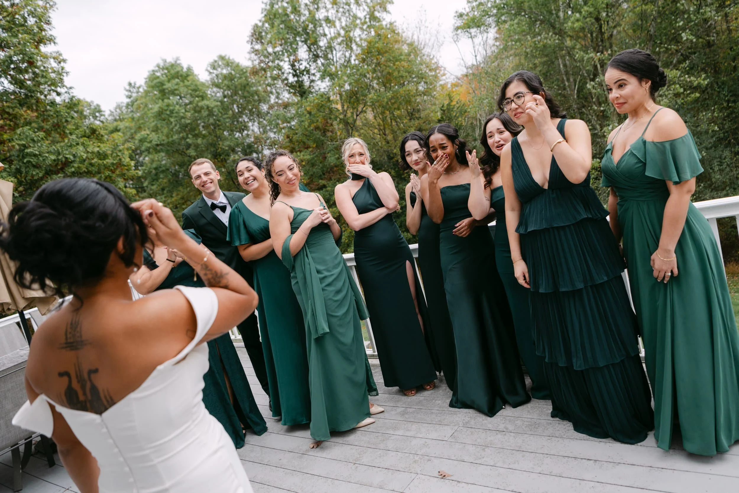 A bride doing a first look with her bridesmaids and groomsmen on an outdoor deck surrounded by trees, all dressed in dark green and black formal attire.
