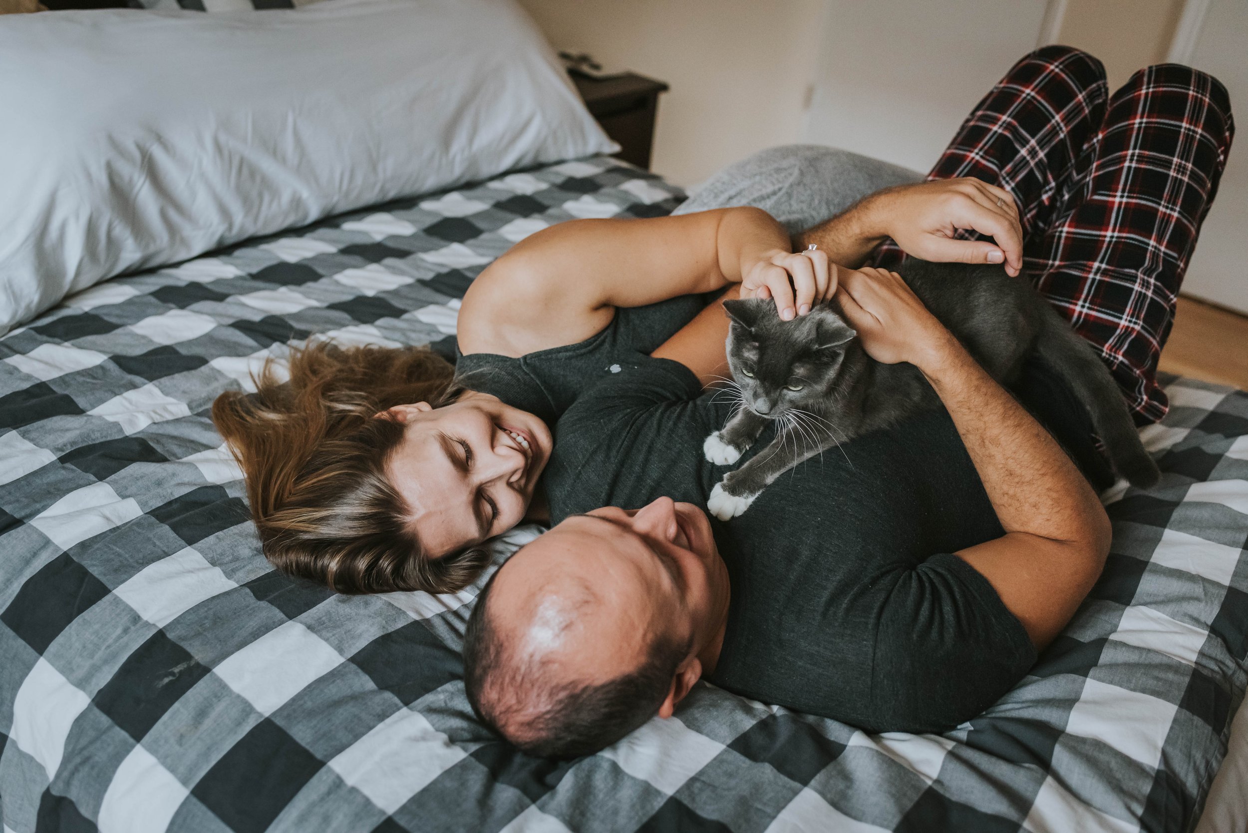 A couple lying on a bed, playing with a gray cat, with both of them smiling and looking at the cat.