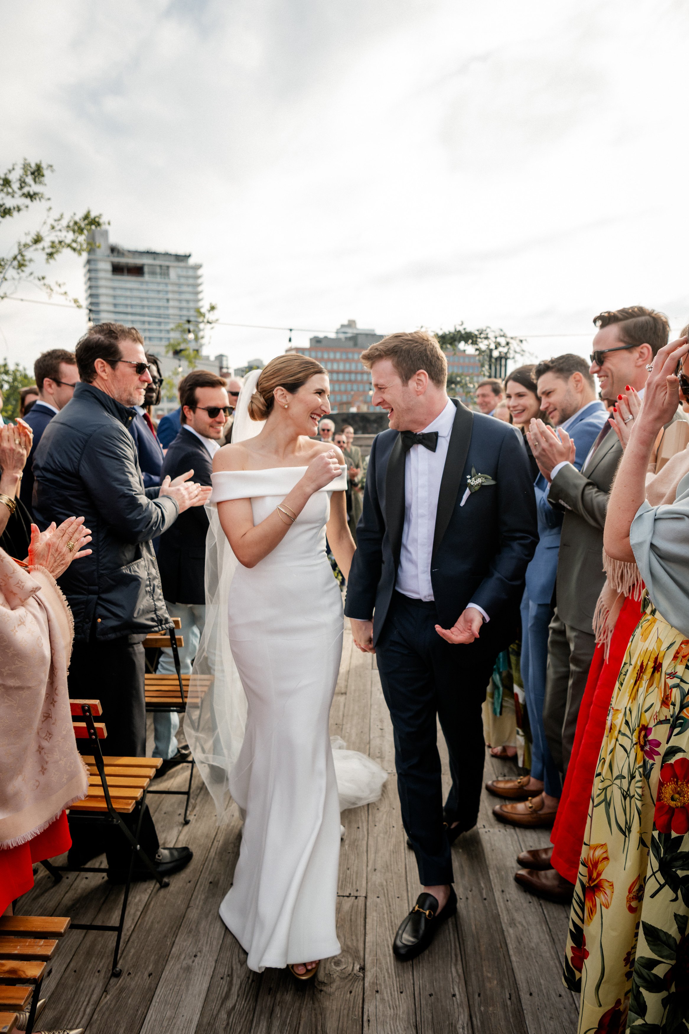 A bride and groom happily walking hand in hand on a wooden deck during their wedding celebration, surrounded by smiling guests clapping and enjoying the moment outdoors with city buildings in the background.