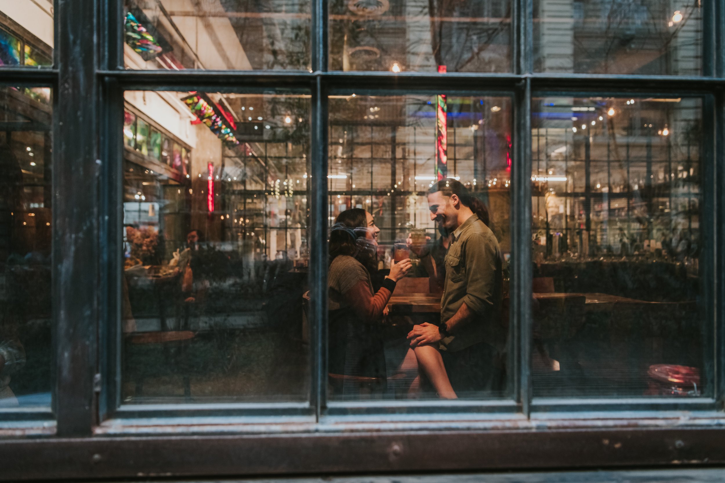 People sitting and enjoying drinks at a bar or restaurant, seen through a large window, with reflections of interior and exterior lights.