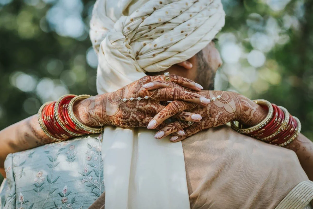 Close-up of a couple embracing, showing the woman's intricately henna-decorated hands with multiple red and gold bangles, resting on the man's shoulder, with green foliage in the background.