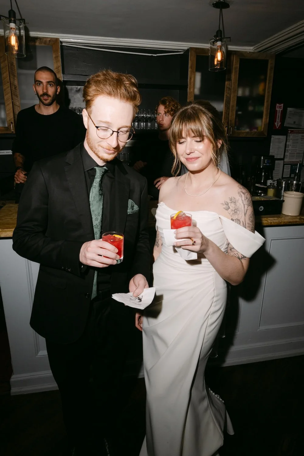 A bride and groom at their wedding reception, holding drinks with lemon slices, standing in a modern bar or restaurant setting, smiling with eyes closed.