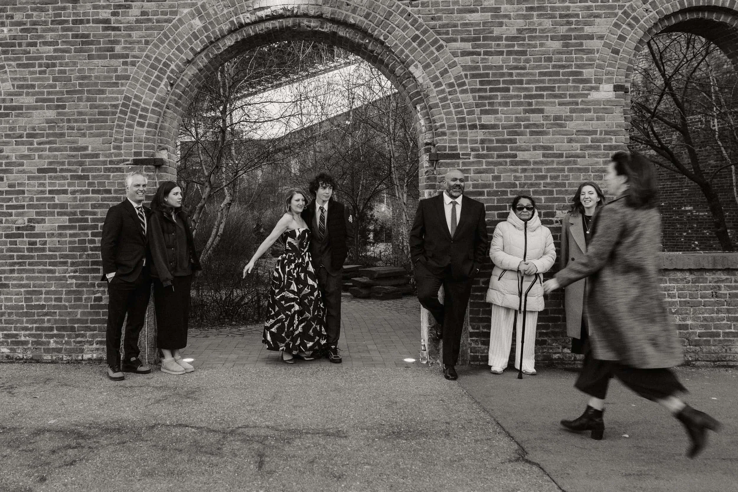 A group of seven people standing by a brick archway outdoors, with a woman walking past in front of them, captured in black and white.