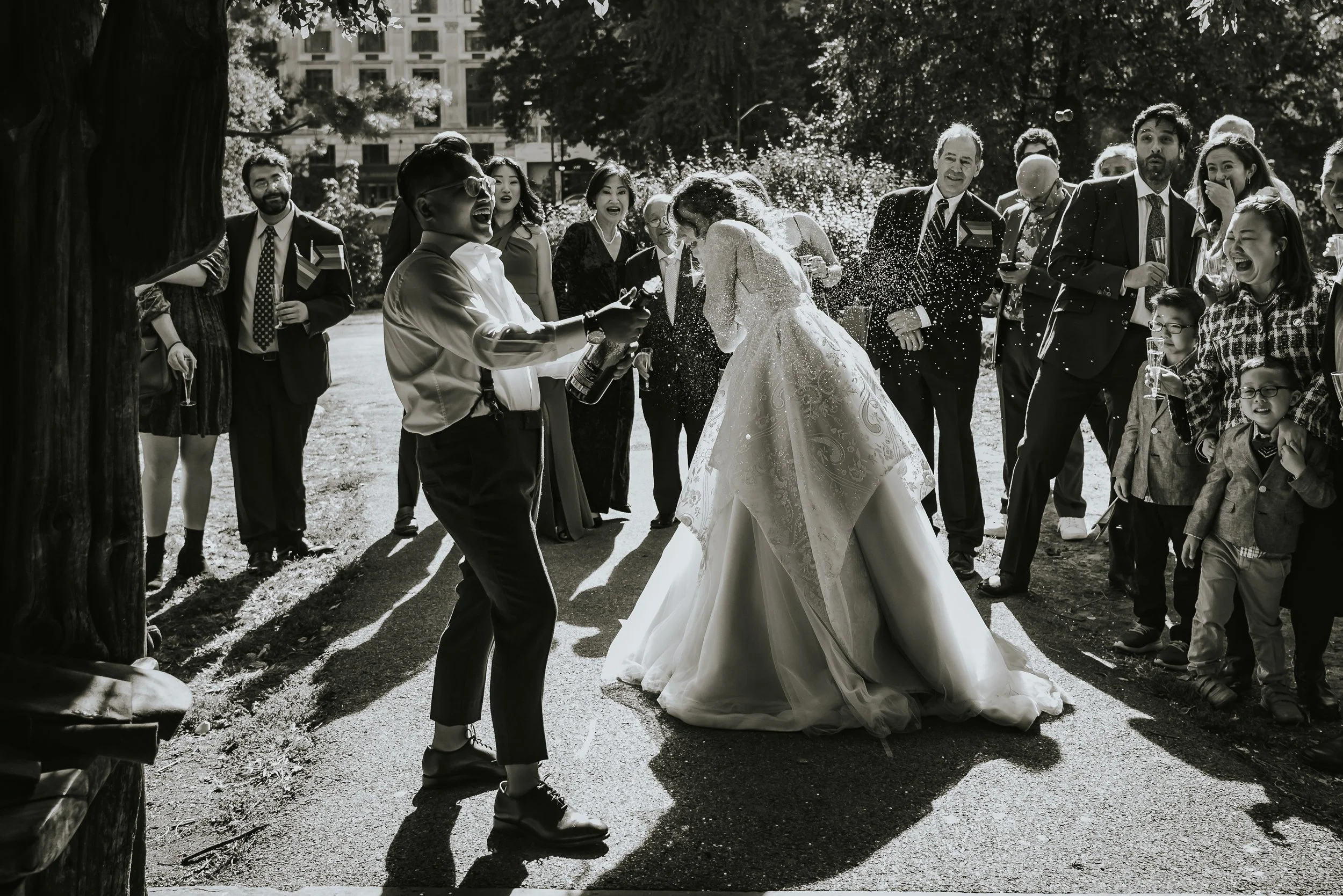 A couple leaning forward in her wedding dress as guests celebrate by spraying her with champagne outside on a sunny day.