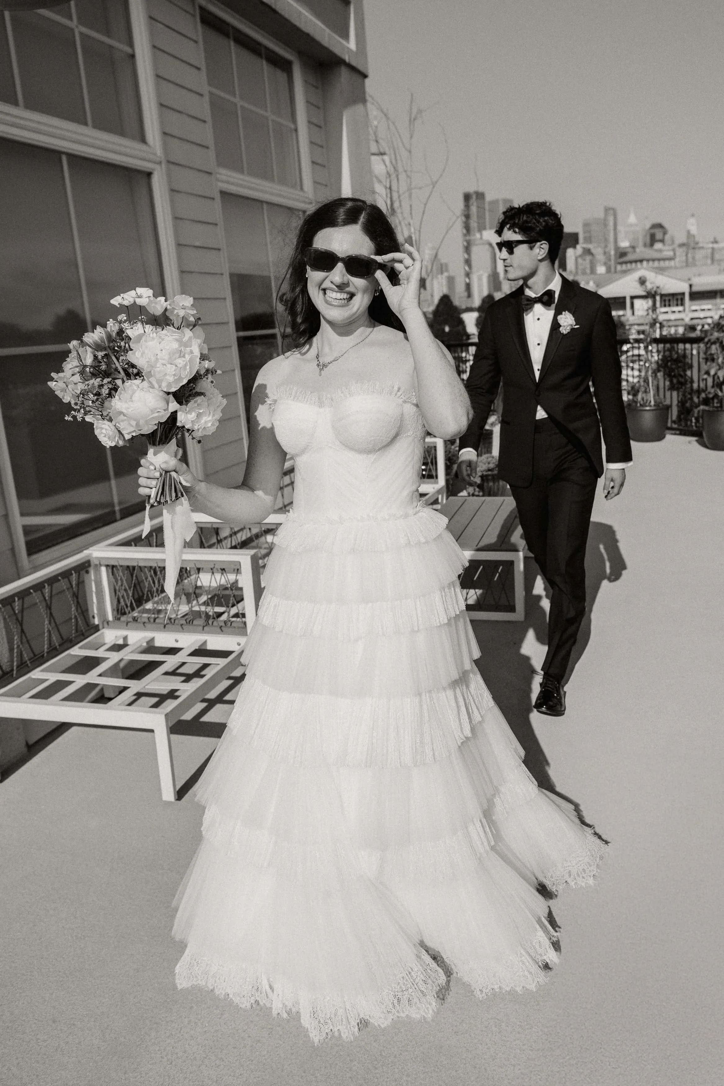 A woman in a wedding dress holding a bouquet of flowers and wearing sunglasses, smiling and adjusting her sunglasses, with a man in a tuxedo walking behind her on a rooftop terrace.