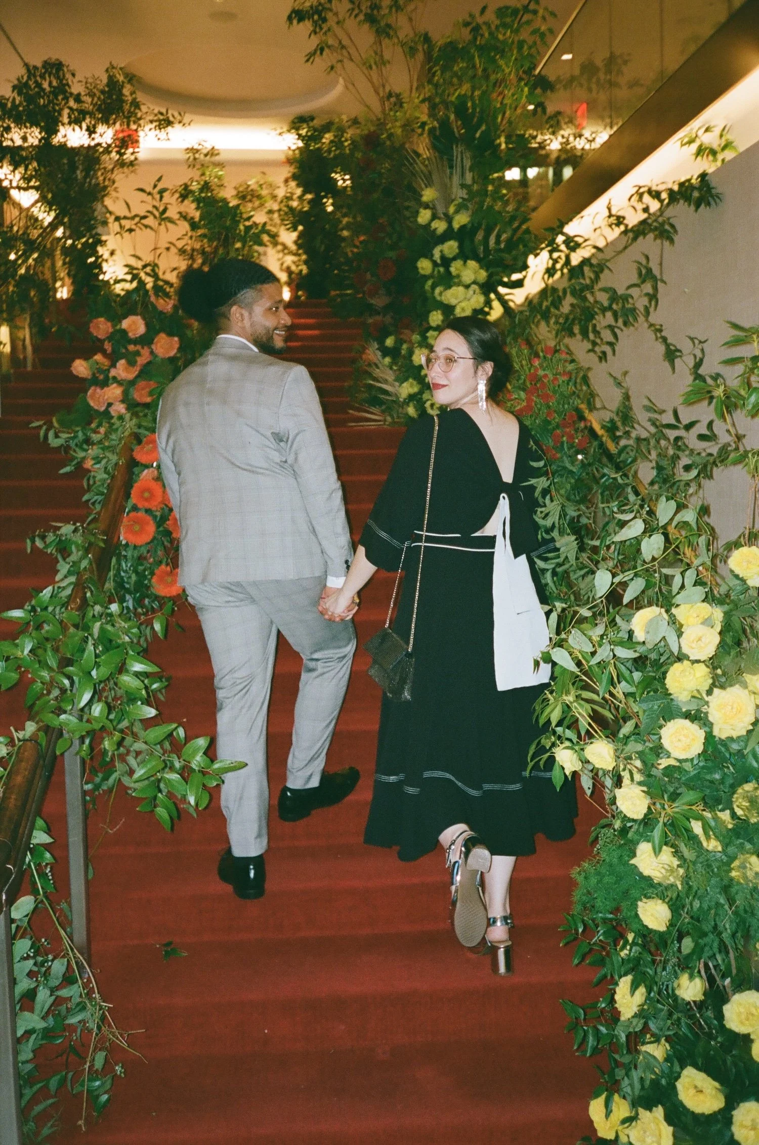 A man and woman holding hands and walking up a red carpeted staircase surrounded by lush green plants and yellow flowers, in an indoor setting.