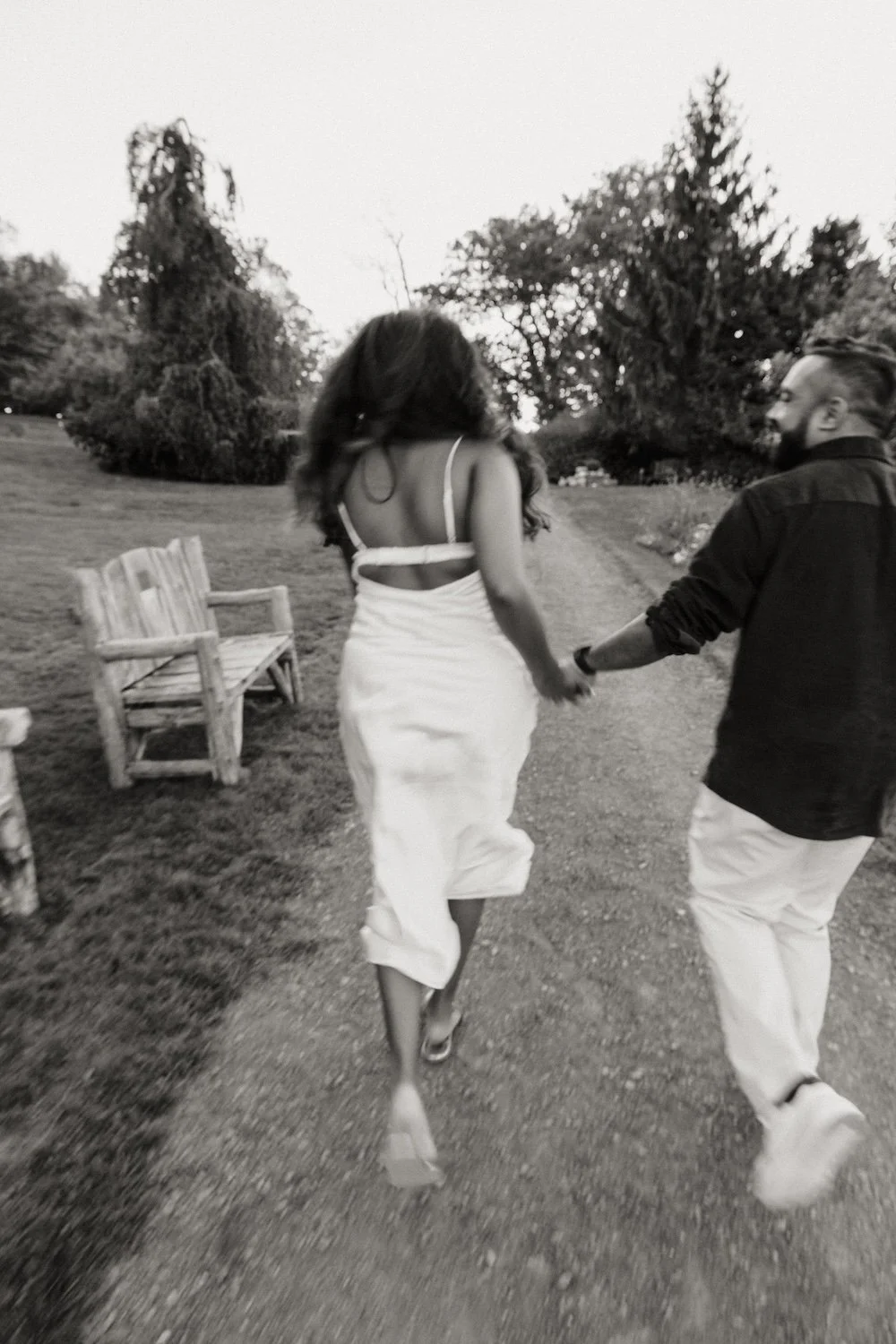 A black and white photo of a couple holding hands and walking outdoors, surrounded by trees and a bench.