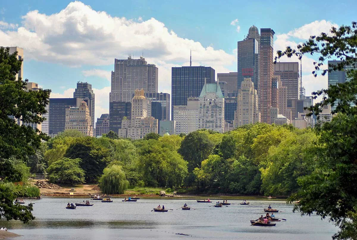 View of a city skyline with tall buildings and a park with a lake and people in small boats