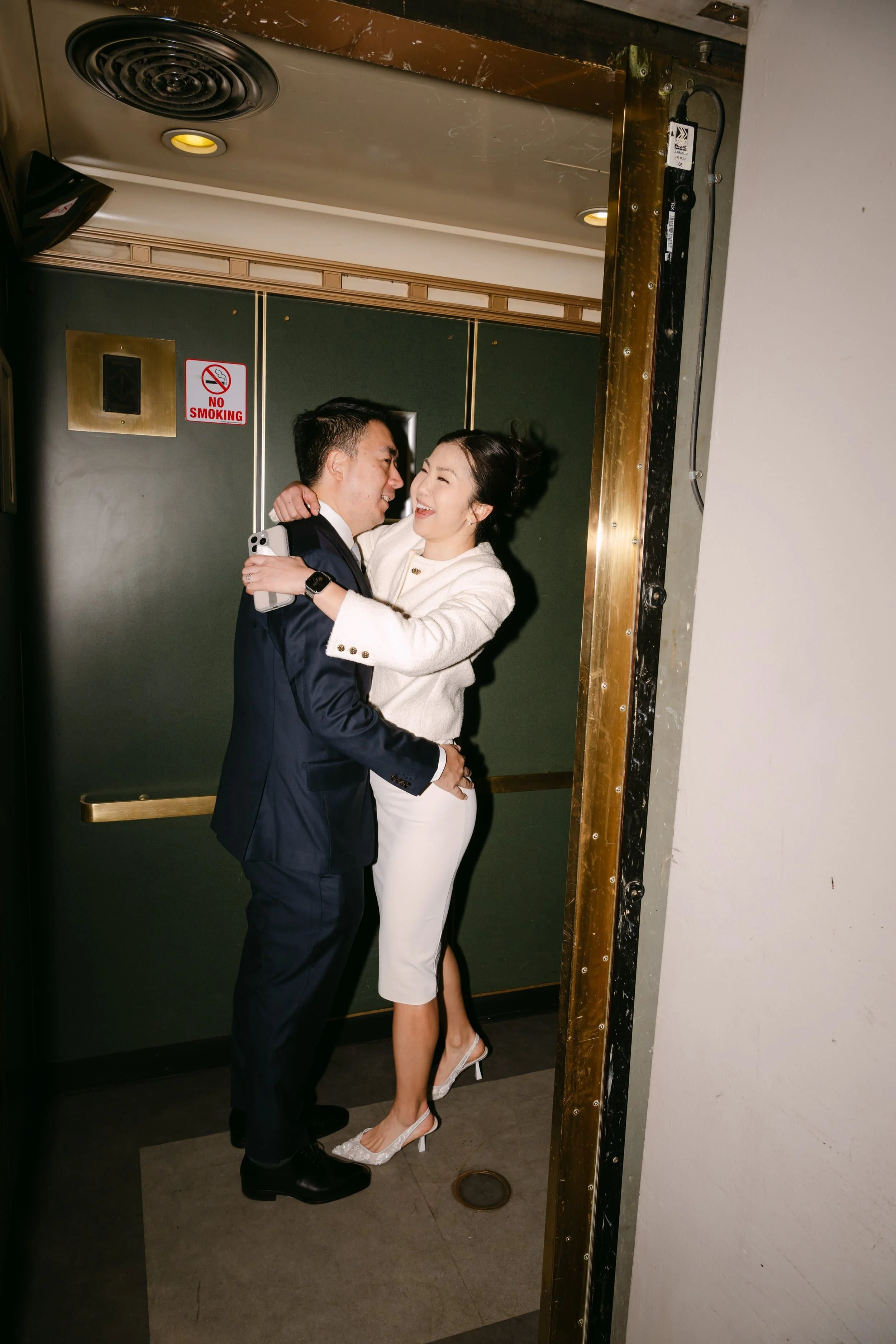 A couple in formal attire embracing happily in an elevator hallway.
