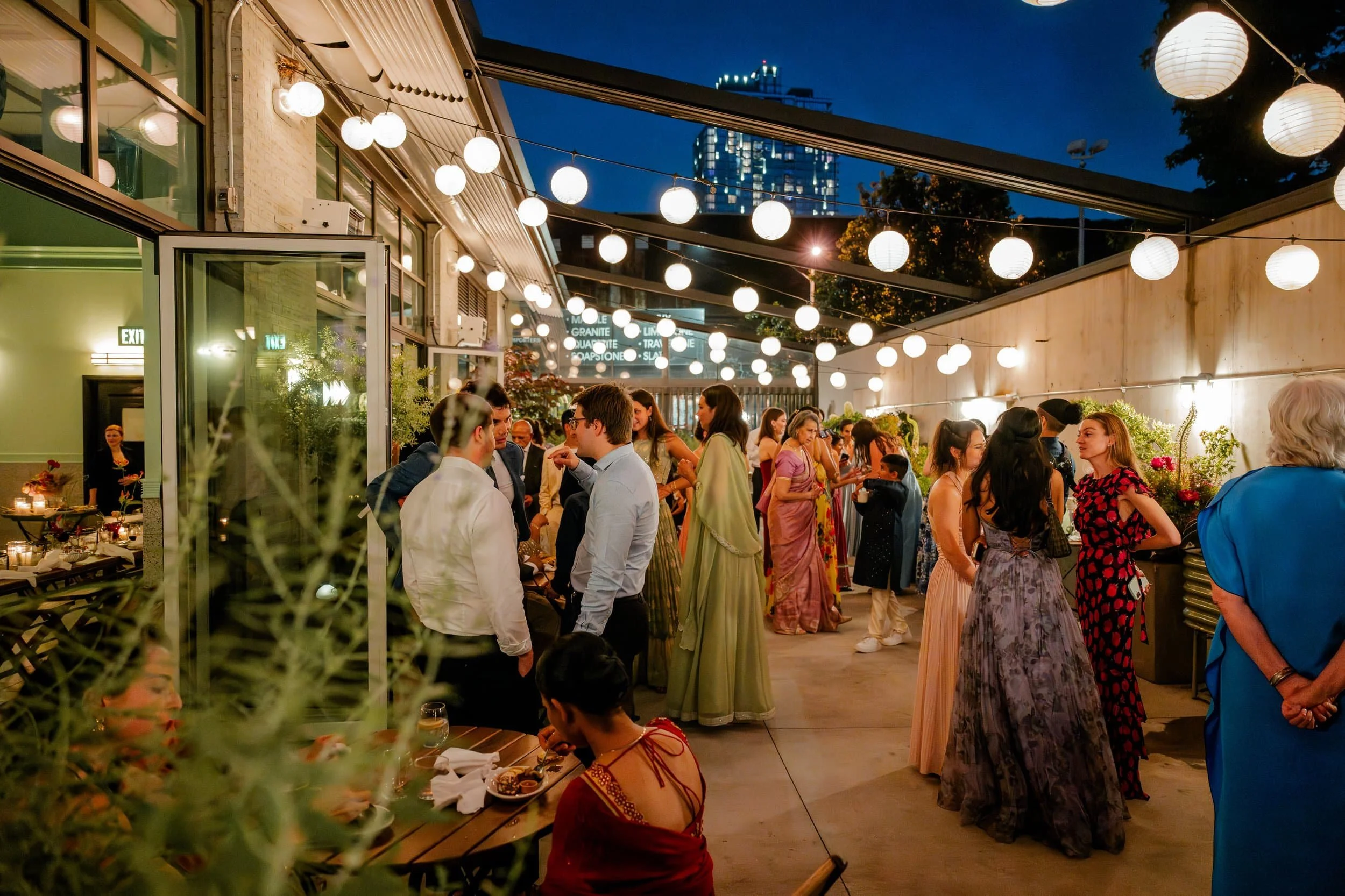 An outdoor evening gathering with people socializing under string lights, with a modern building and city skyline in the background, and decorated with flowers.