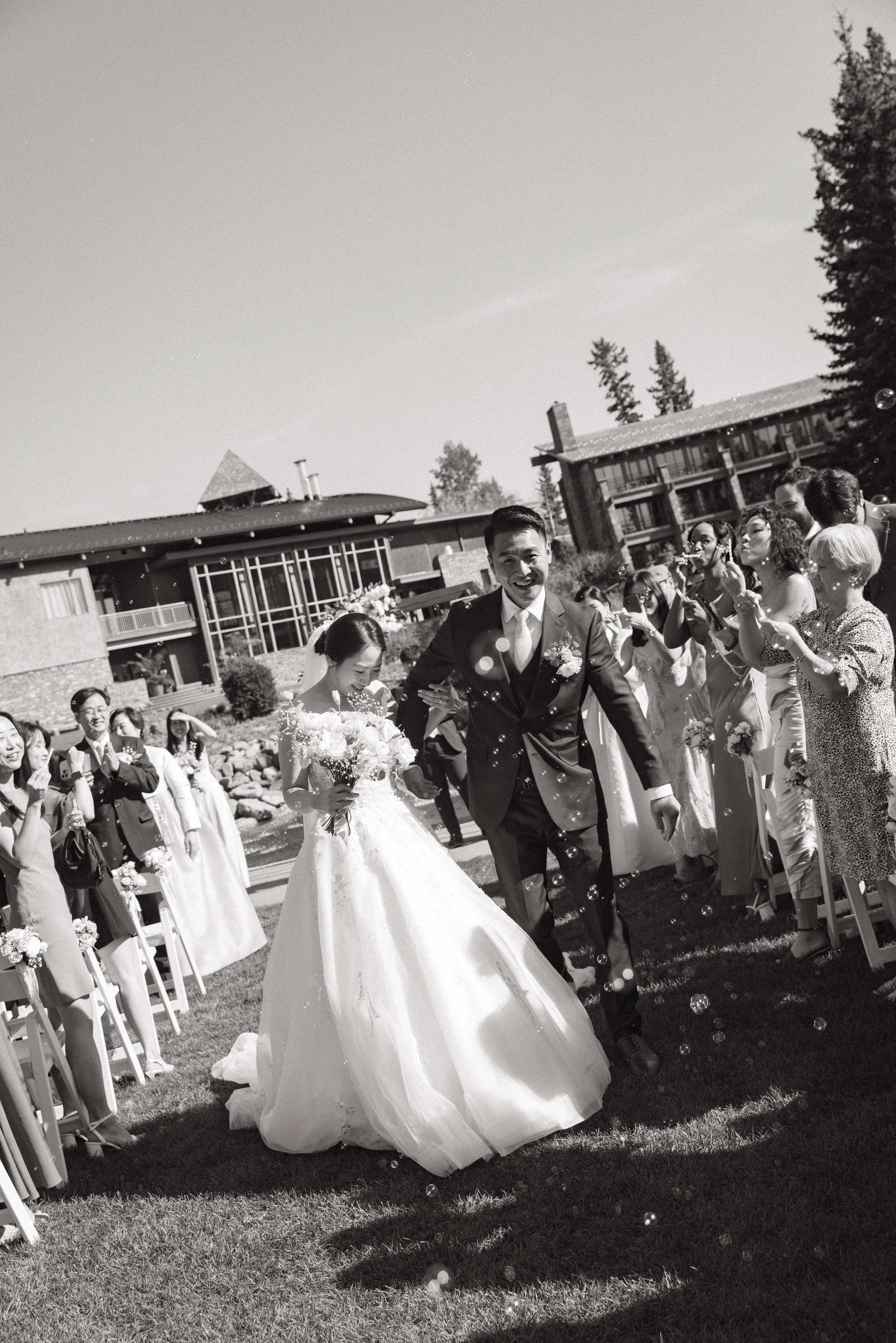 A black and white photo of a wedding scene outdoors. The bride and groom are walking down the aisle, with the bride holding a bouquet and the groom smiling. Guests are standing on either side, smiling and holding sparklers. In the background, there a