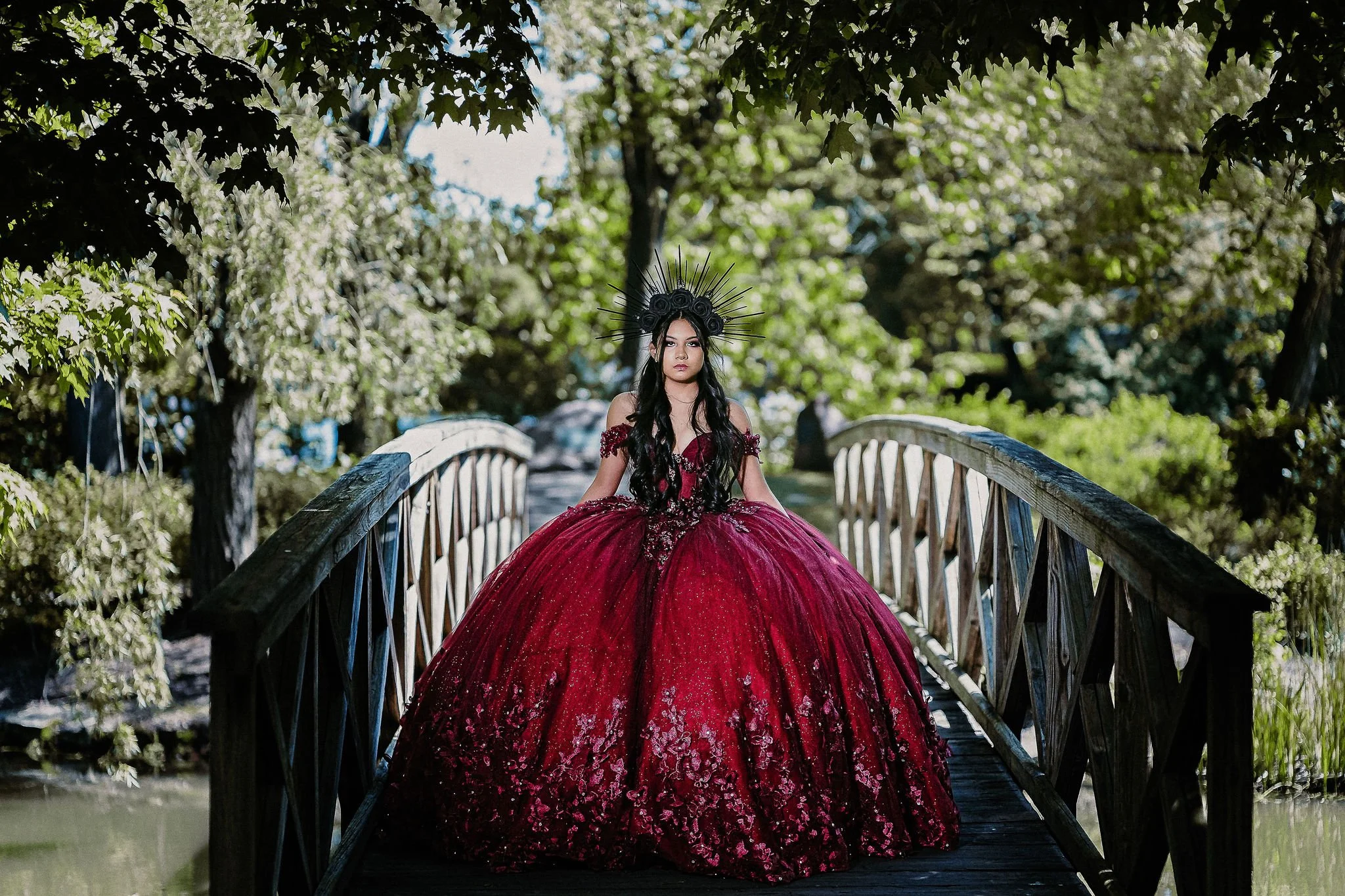 Quinceañera in a dramatic red ball gown with a gothic enchanted forest theme on a garden bridge.