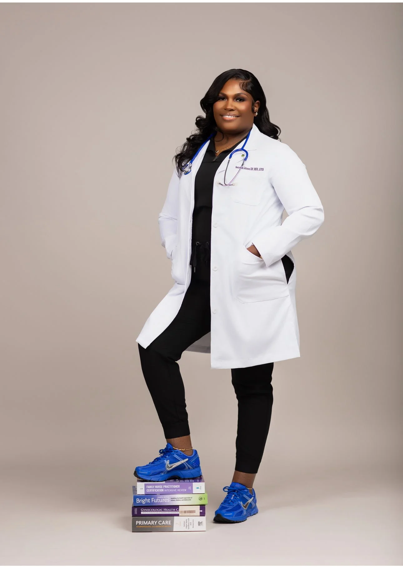 Woman in black scrubs and white lab coat with stethoscope, standing with hand in pocket, five medical books stacked on floor