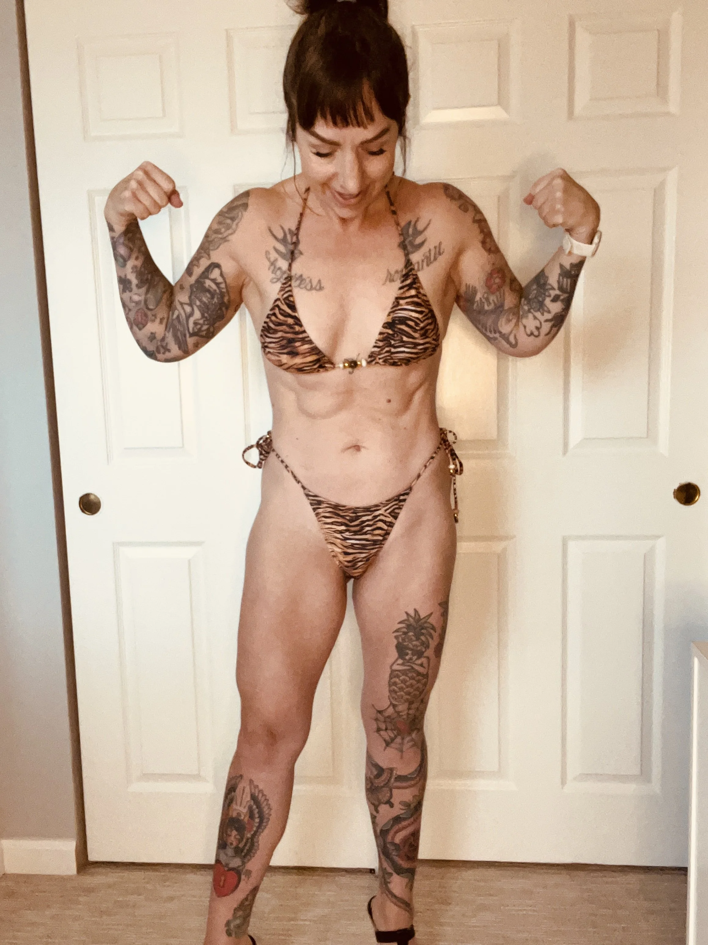 Woman in a tiger-striped bikini flexing her biceps, showing off her tattoos, standing in front of a closed door.