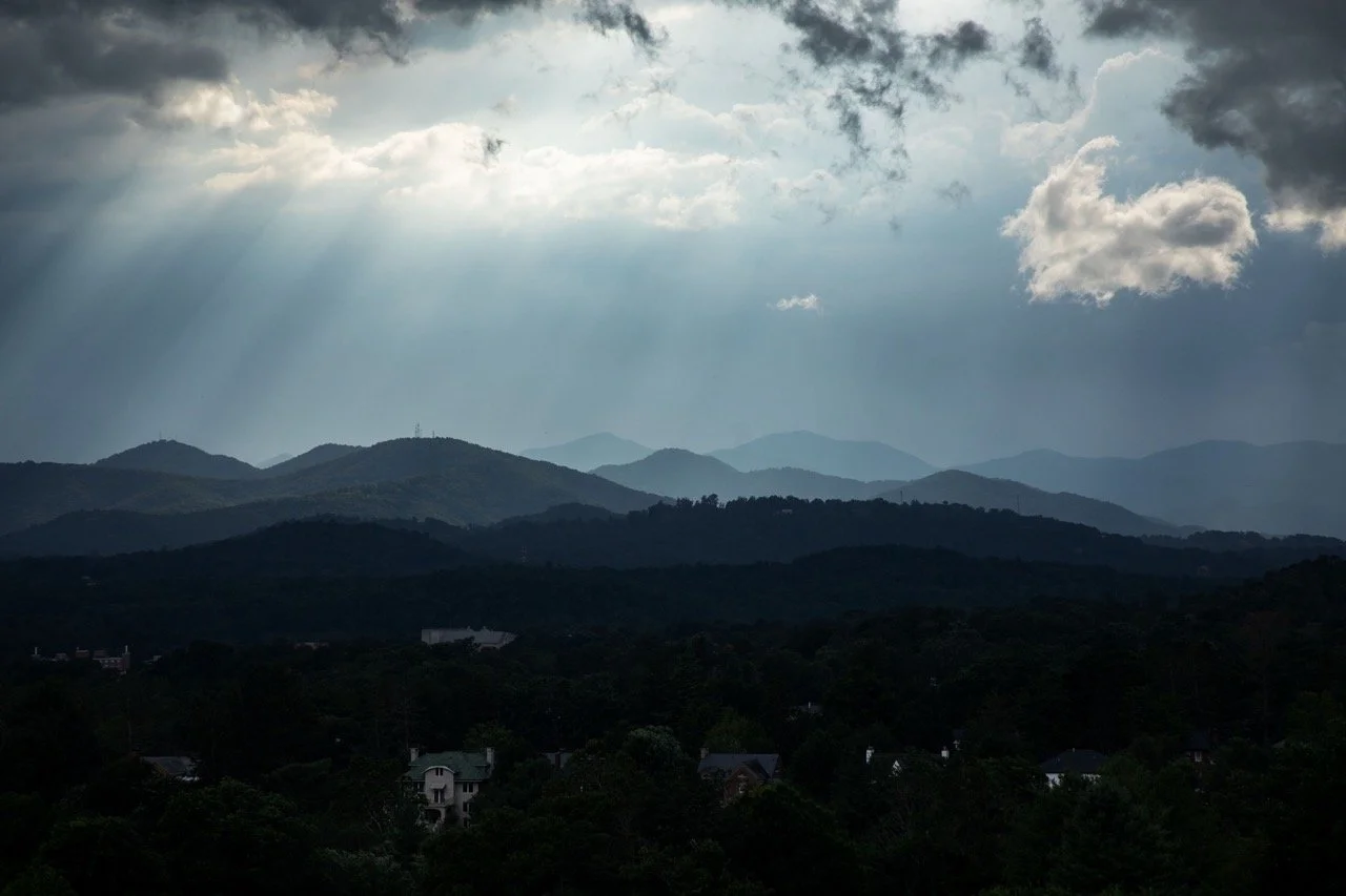 Scenic view of multiple mountain ranges under a cloudy sky with sunlight rays breaking through clouds, overlooking a small town or neighborhood at the valley with trees and houses.