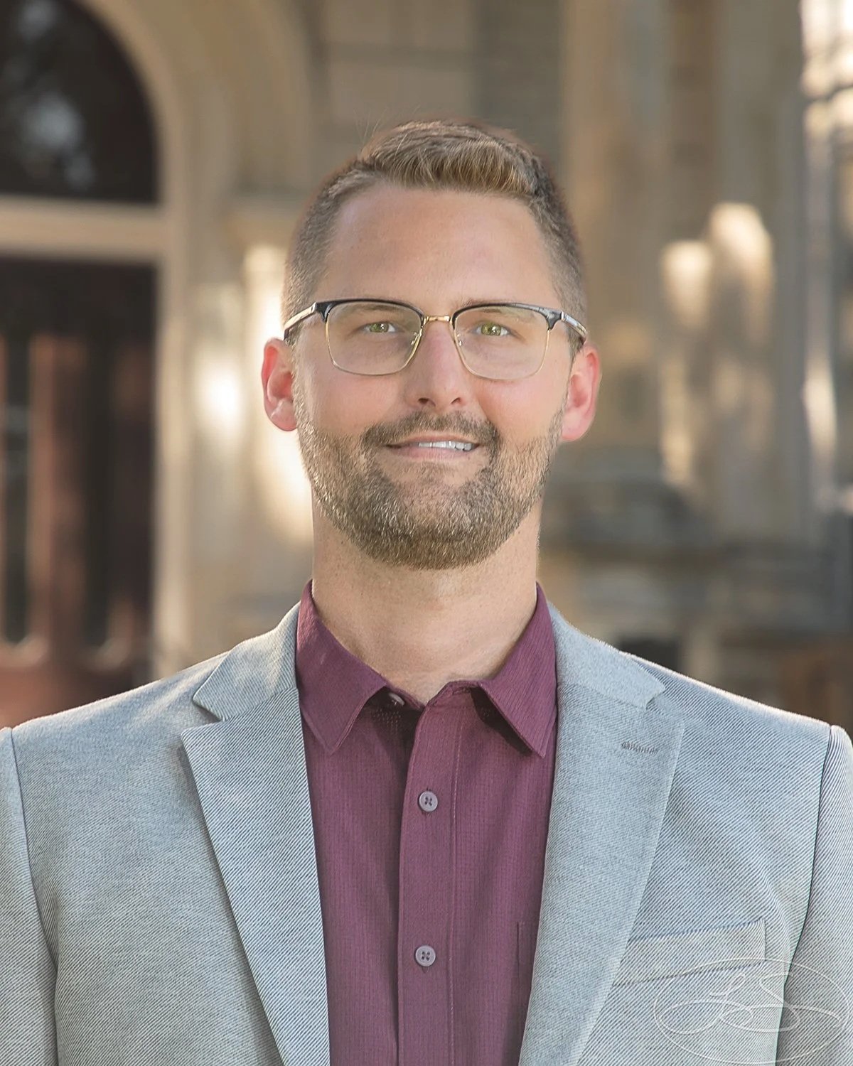 Portrait of a man with glasses and a beard, wearing a light gray blazer and a maroon shirt, standing outdoors in front of a historic building with arched windows and stone architecture.