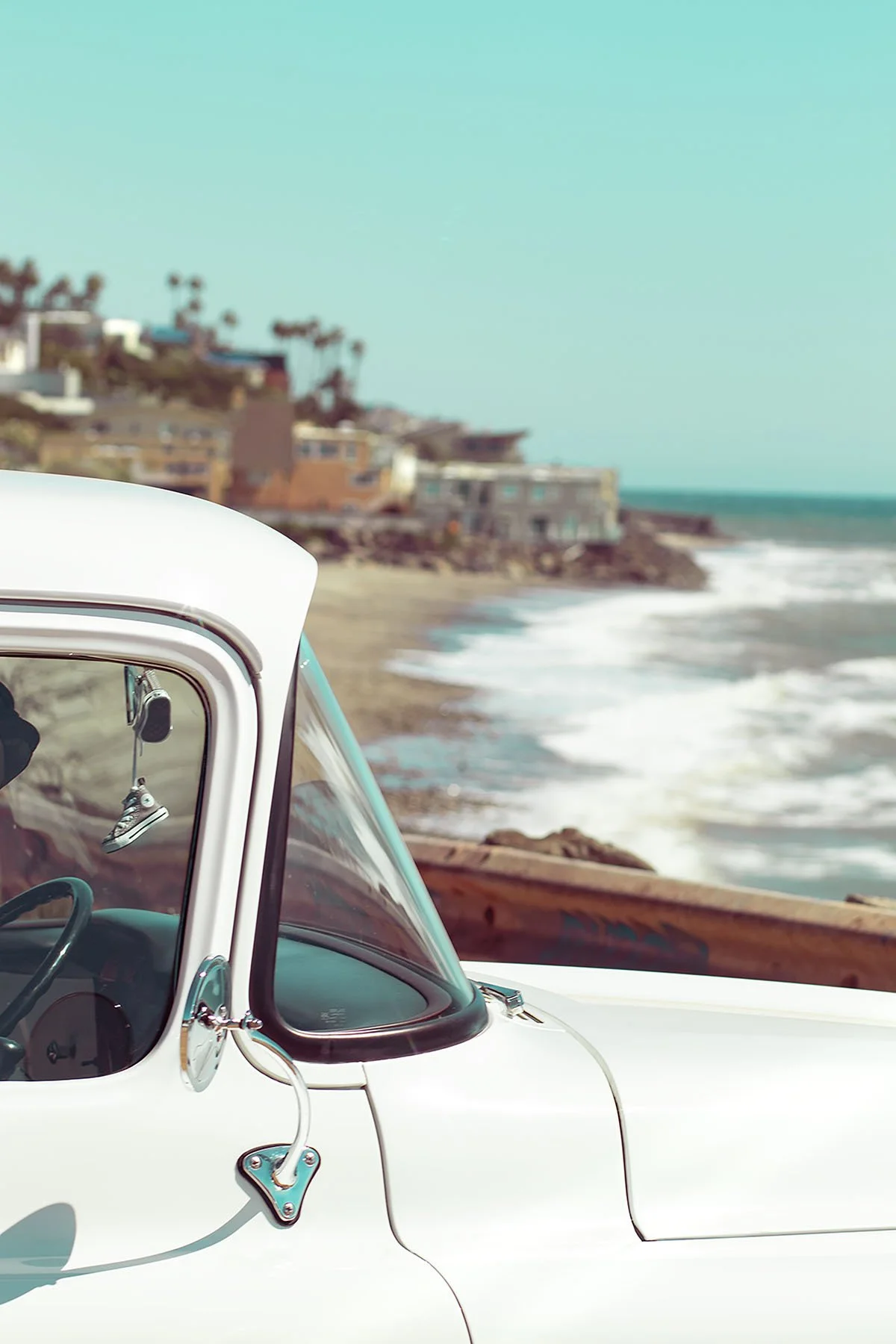 Close-up of a vintage white car parked near a beach with ocean waves and houses in the background.