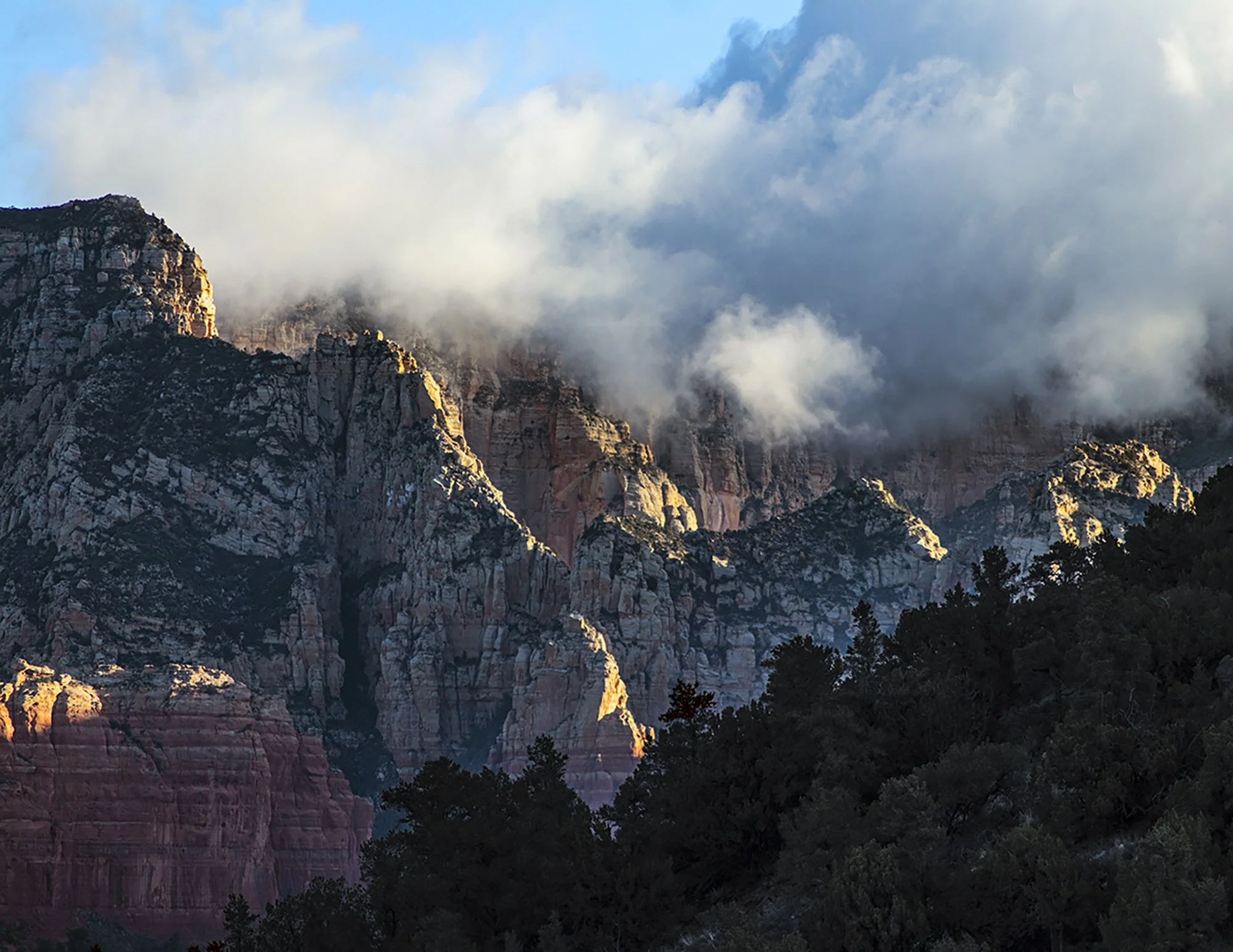 Majestic mountains with rugged, colorful rock formations partially covered by clouds, and dark silhouette of trees in the foreground.