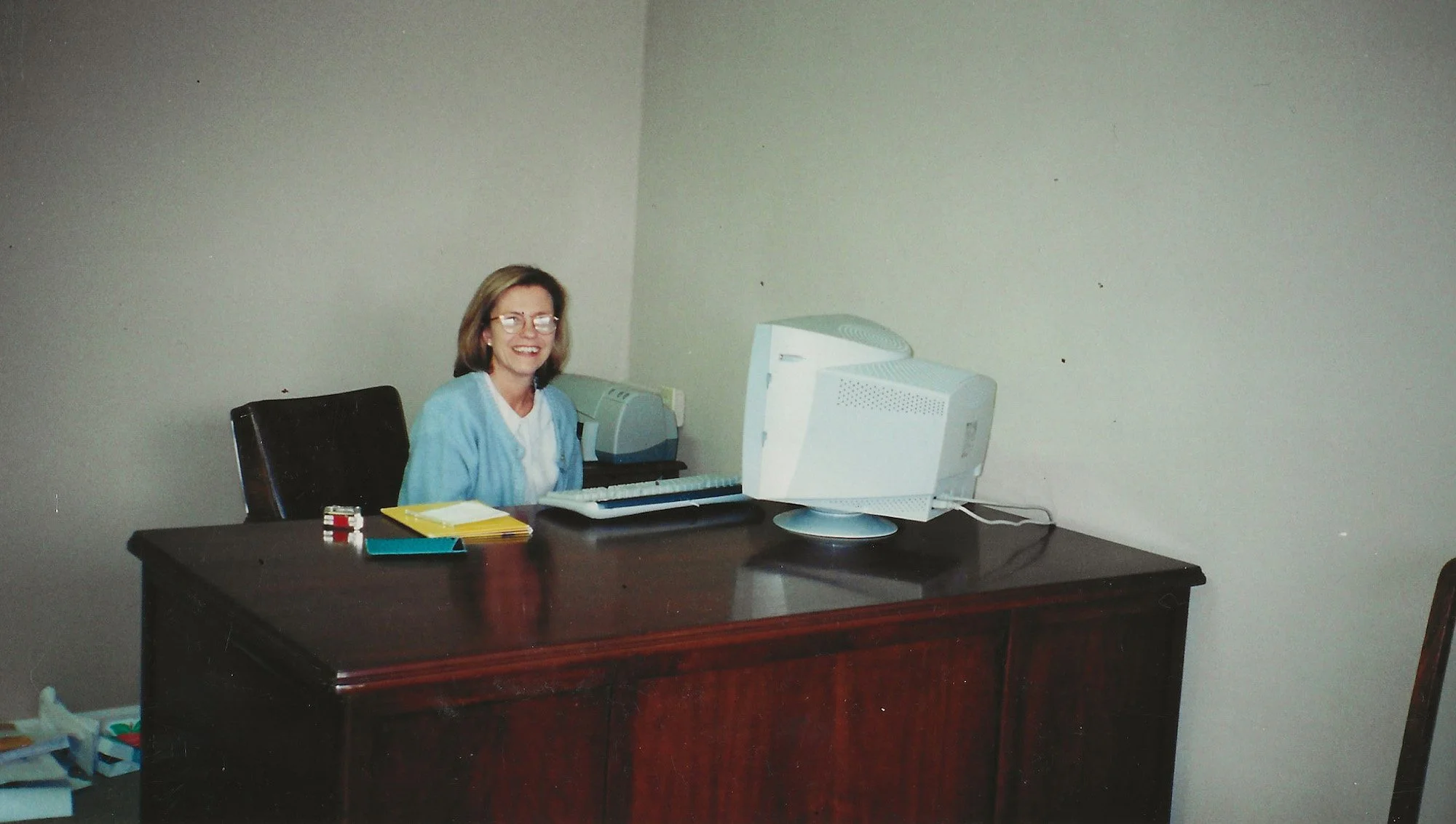 Person sitting at a wooden desk with a vintage computer monitor, smiling.