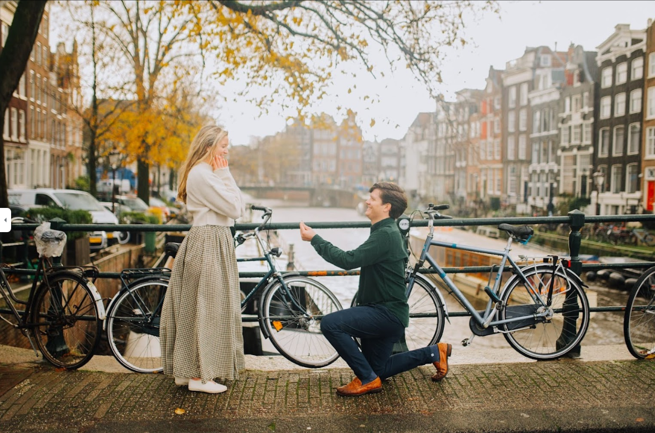 Man proposing to a woman on a canal bridge with bicycles and autumn trees in the background.