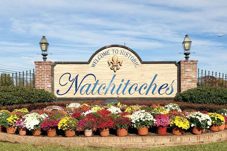 A welcome sign for Historic Natchitoches surrounded by colorful flowers and two brick columns with lamps.