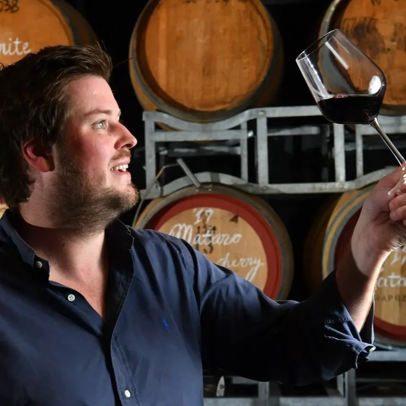 A man holding a glass of red wine in a wine cellar with barrels in the background.