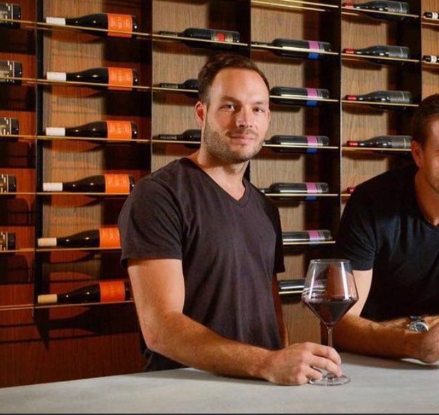 A man holding a glass of red wine while standing at a wine tasting bar, with wine bottles displayed on shelves behind him.