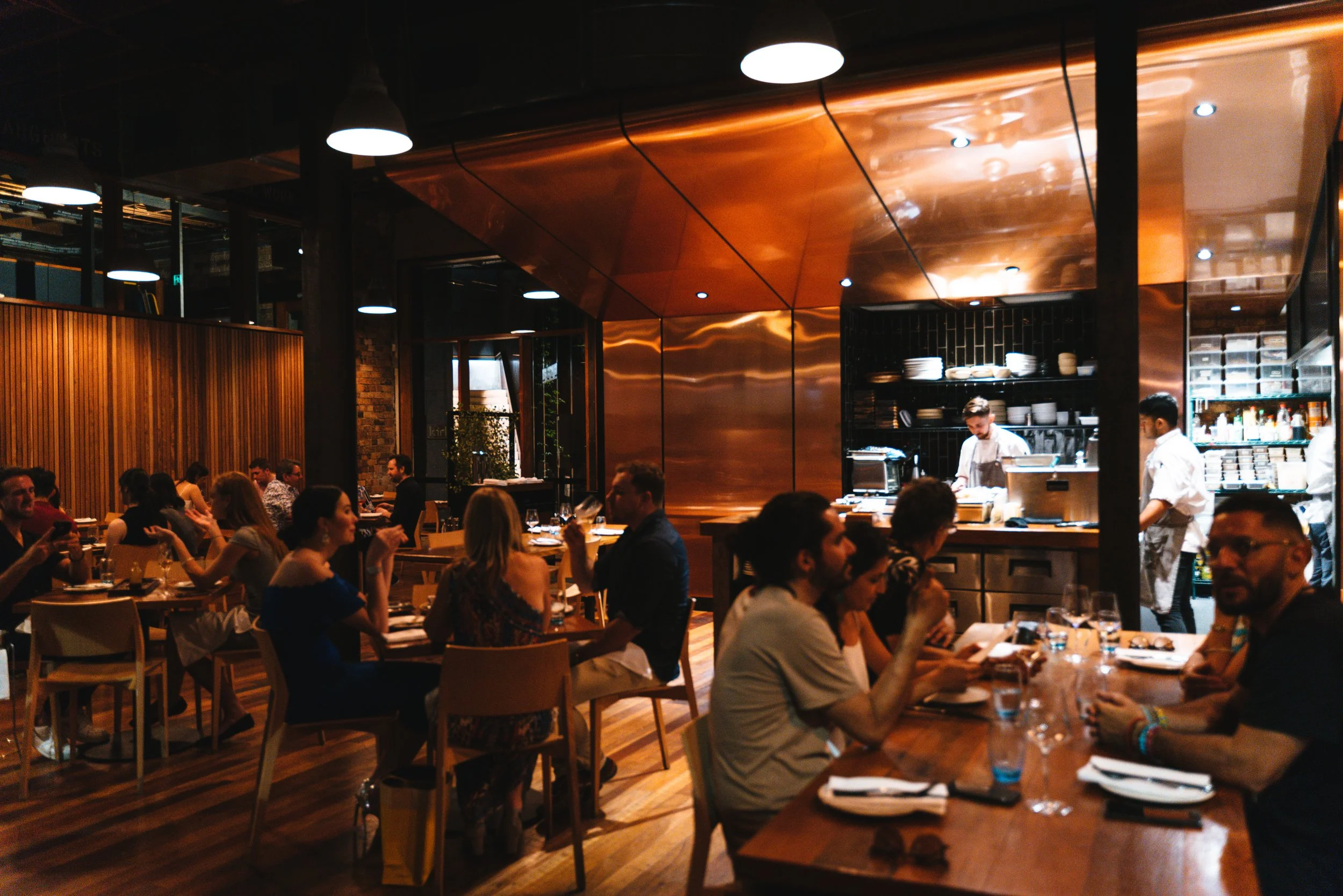 Interior of a lively restaurant with patrons dining at tables, chefs working in an open kitchen, warm lighting, wooden accents, and a modern design.