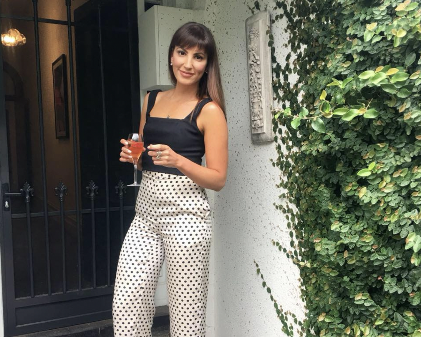A woman in a black sleeveless top and white polka-dot pants holding a glass of pink drink, standing outside near a white wall with green foliage.