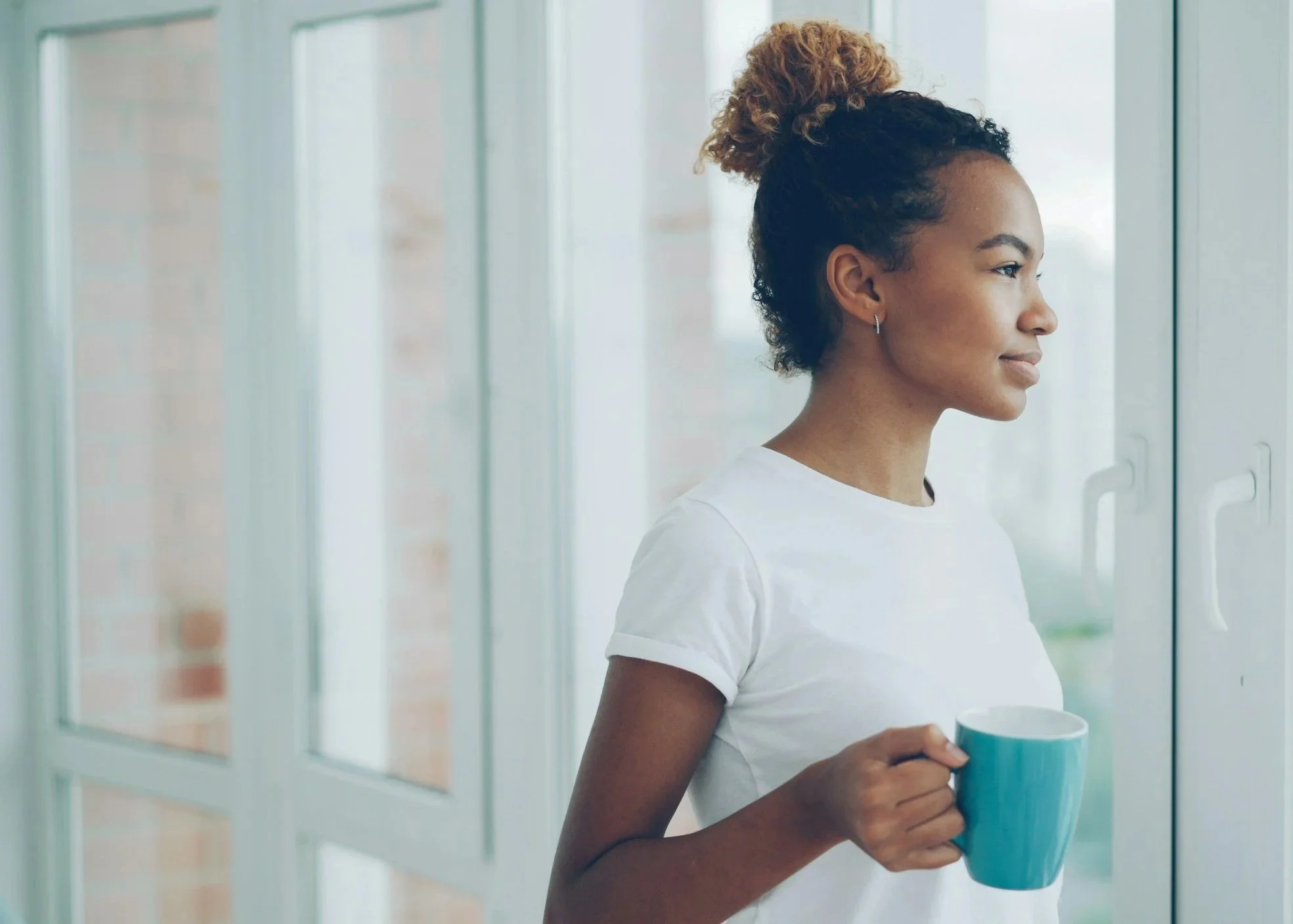 Profile of a young Black woman with curly hair tied up in a bun, wearing a white t-shirt, holding a blue mug, standing near glass doors in a bright indoor setting.