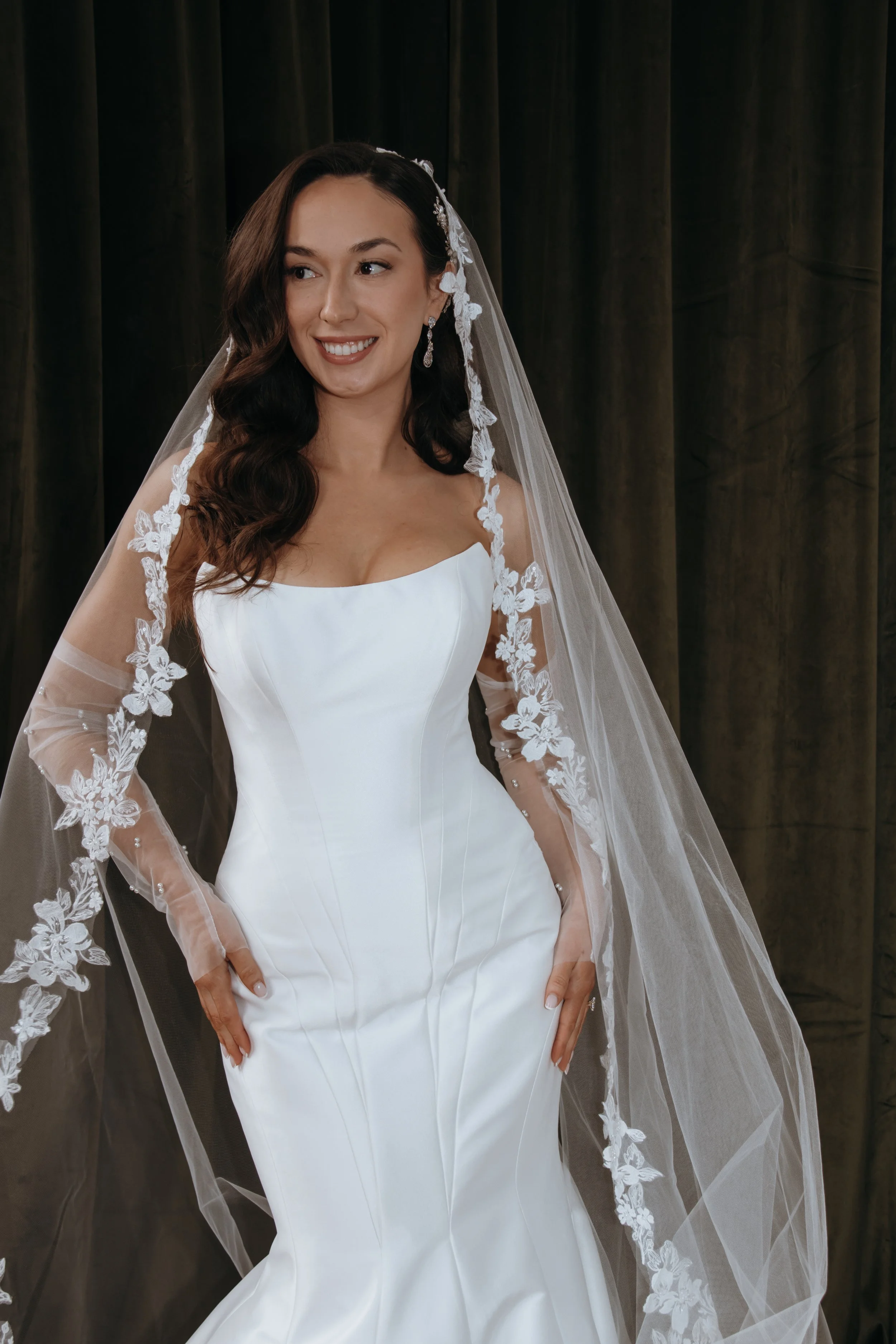 A woman in a white wedding dress with a veil, standing in front of a dark curtain, smiling. Makeup by Georgia Lipscombe - Georgia Peach Beauty photographer Anna Lawton @photographyby.anna