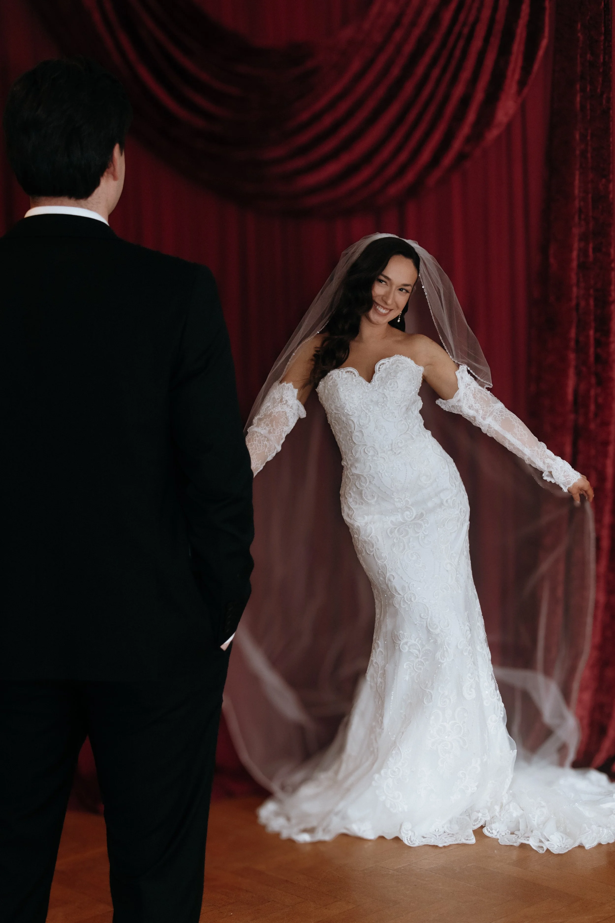 A bride in a white lace wedding gown and veil is smiling at a groom in a black suit during their wedding ceremony in front of red curtains. Makeup by Georgia Lipscombe - Georgia Peach Beauty photographer Anna Lawton @photographyby.anna
