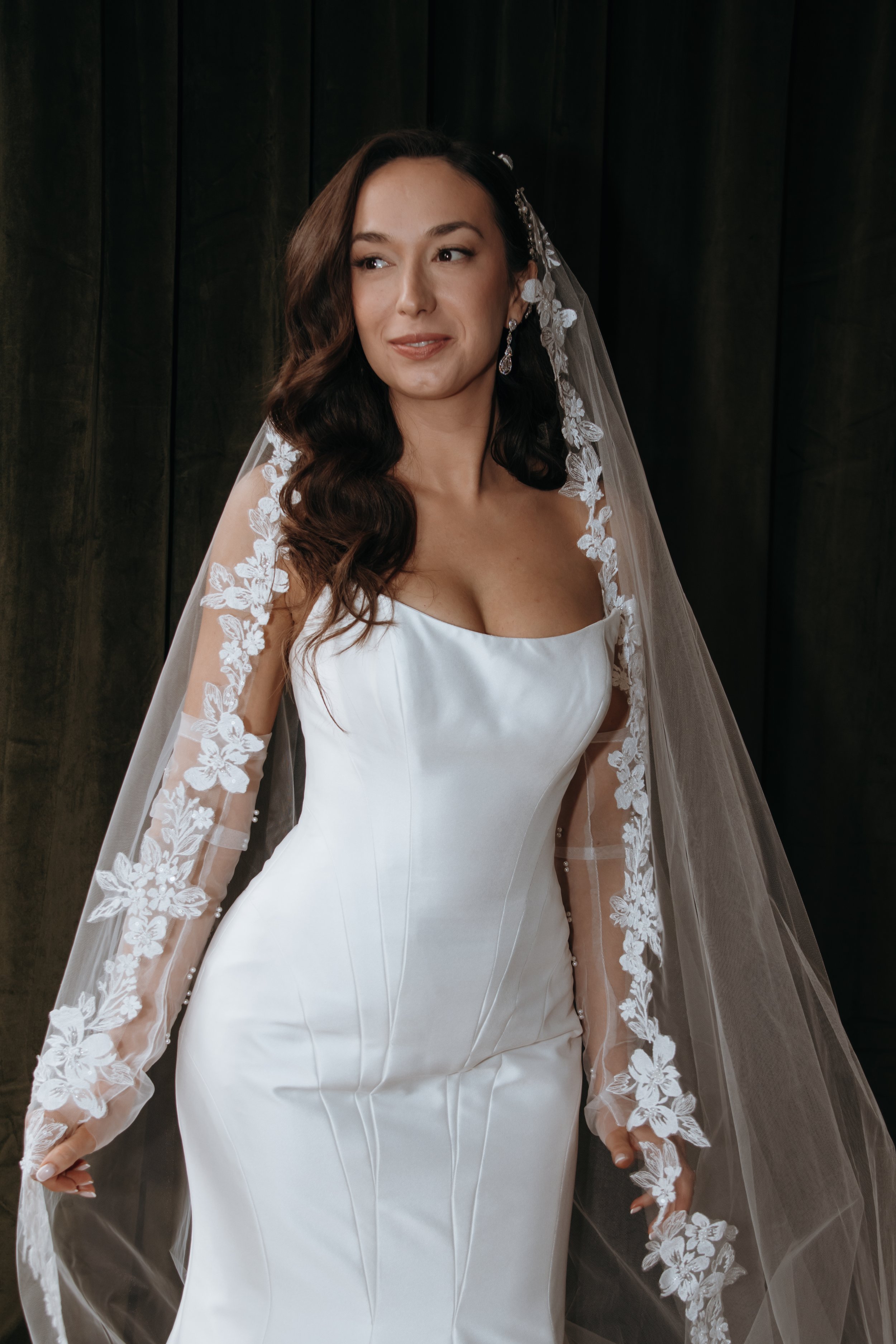 Woman wearing a white wedding dress with lace sleeves and a floral lace veil. She has long, dark wavy hair and is standing in front of a dark background. Makeup by Georgia Lipscombe - Georgia Peach Beauty photographer Anna Lawton @photographyby.anna