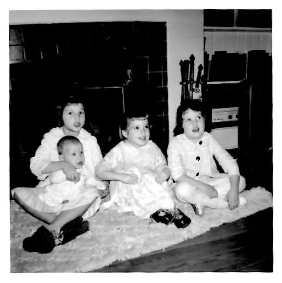 Young Julia Brim-Edwards with siblings sitting on a carpet in a living room, with a woman behind them in the background. The children are all wearing light-colored dresses and appear to be happy and smiling.