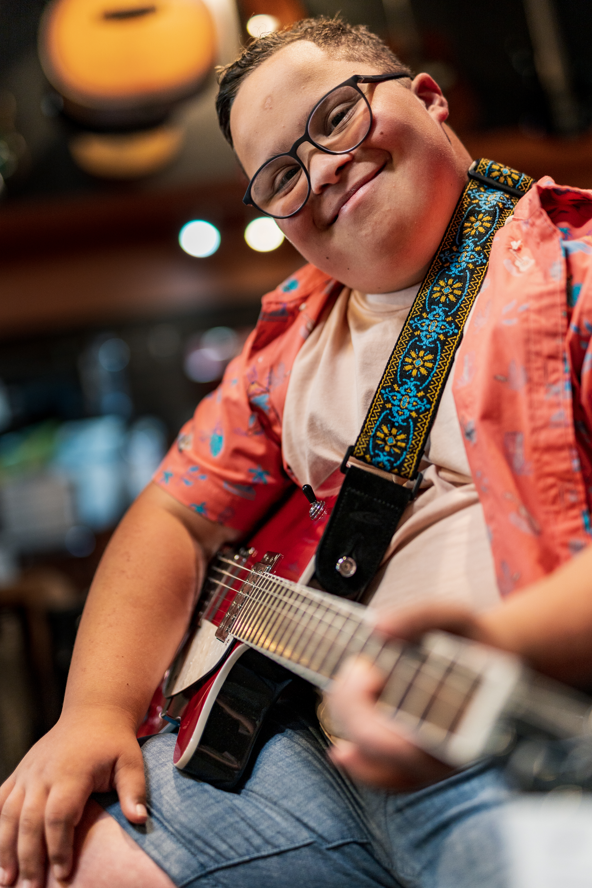 A young person with glasses smiling and playing an electric guitar, wearing a patterned strap, a flower-patterned shirt over a beige t-shirt, and shorts, in a lively indoor setting.
