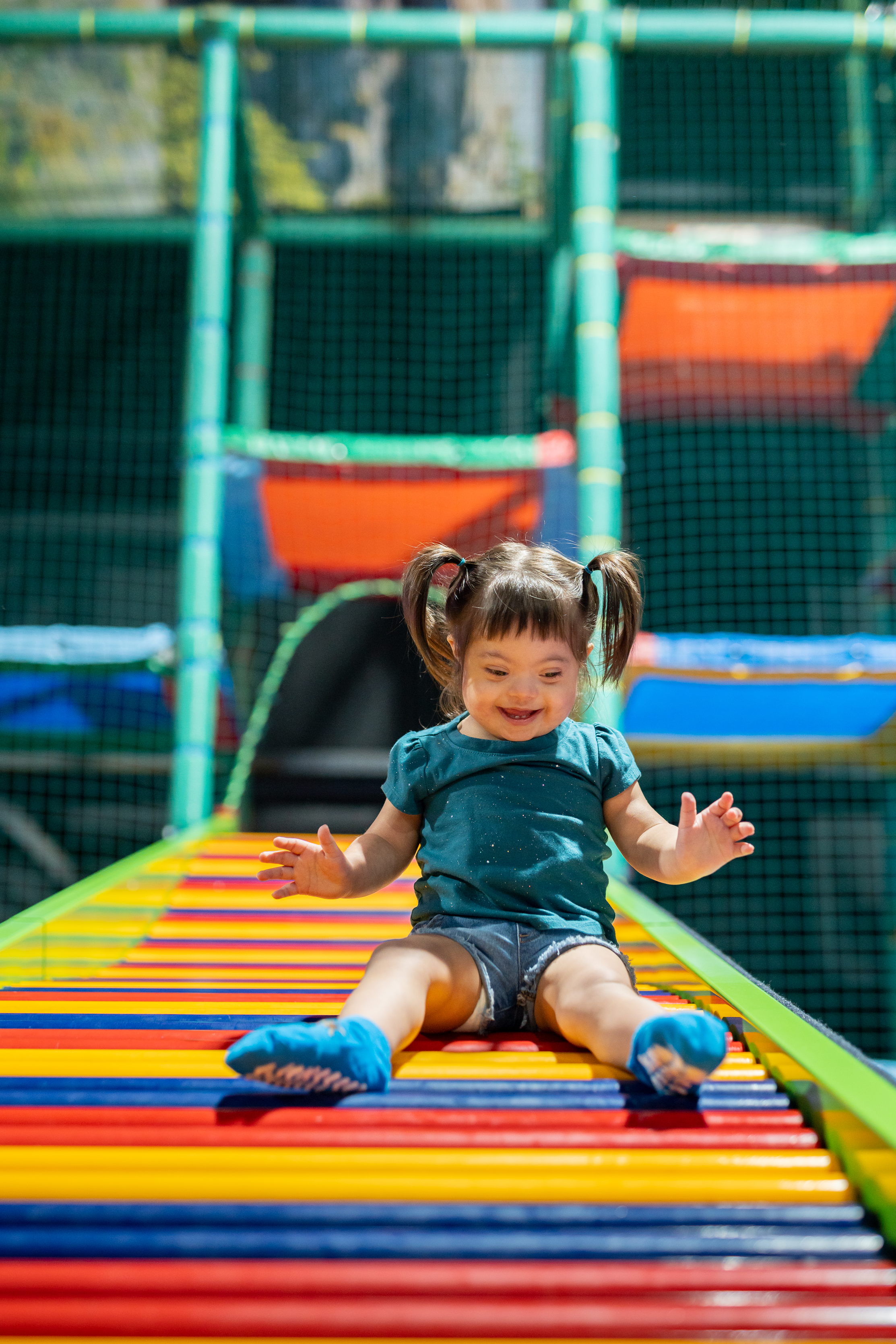 A young girl with pigtails sliding down a colorful play structure at an indoor playground, surrounded by safety netting.
