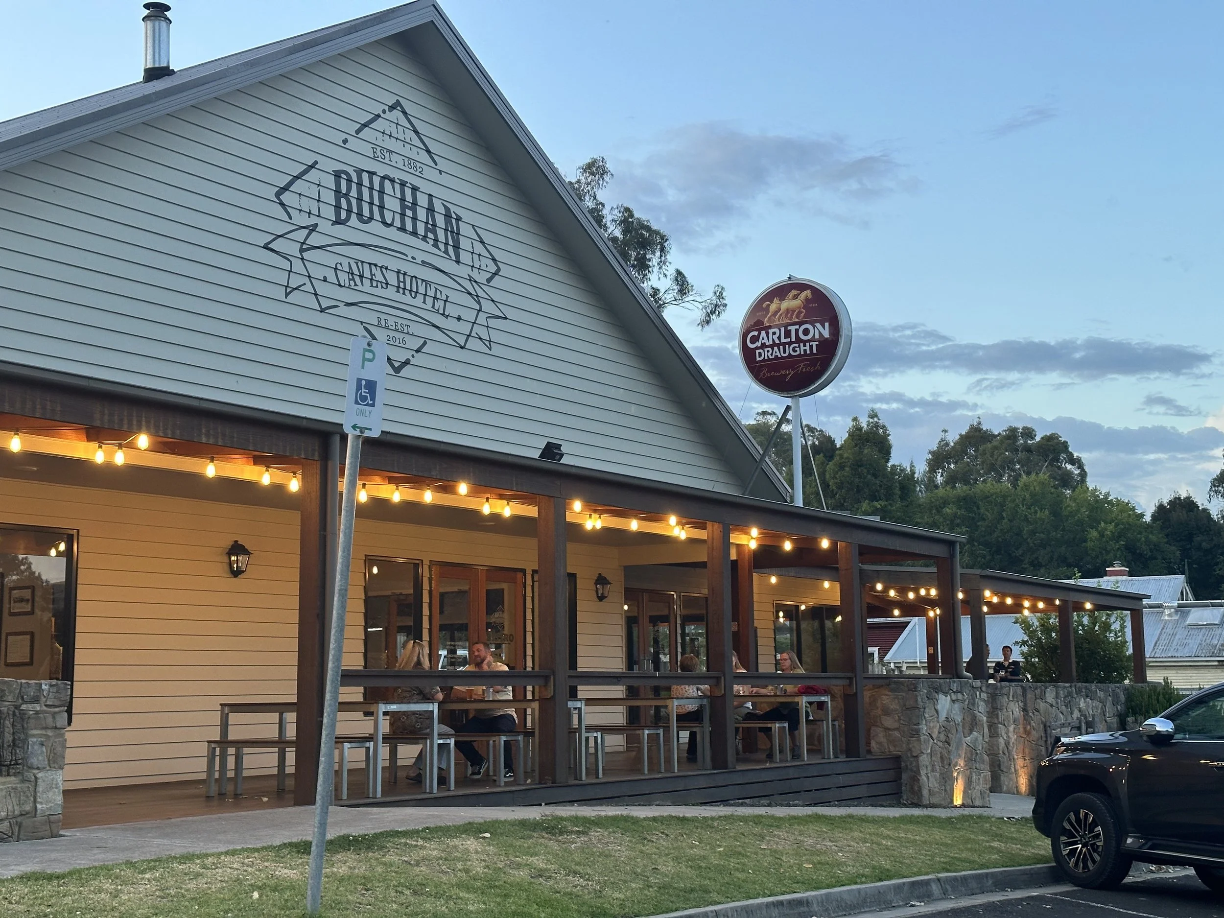 Exterior view of Buchan Caves Hotel with outdoor seating area illuminated by string lights, a Carlton Draught sign in the background.