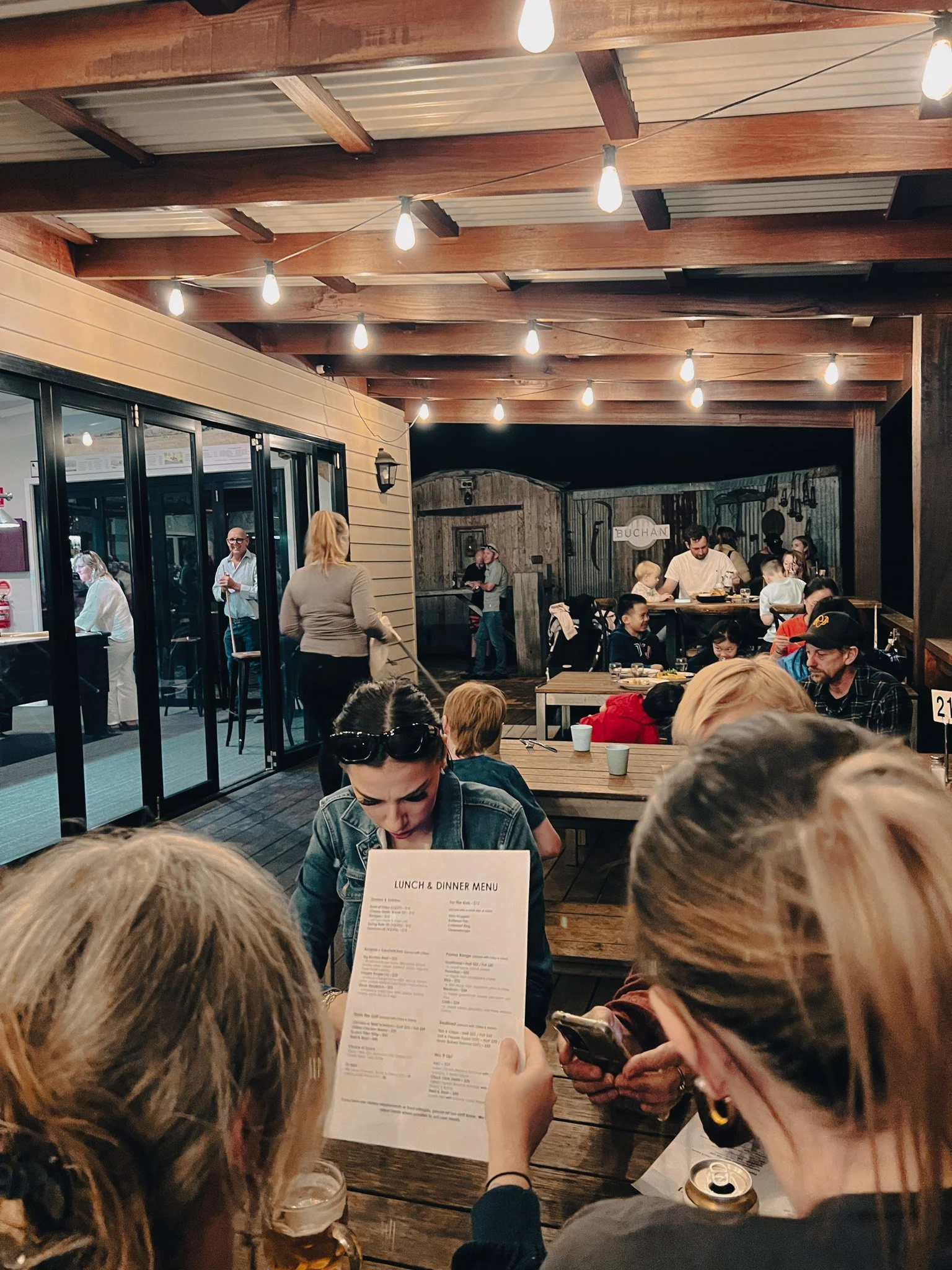 People sitting at a wooden table a Buchan Caves Hotel with string lights hanging from wooden ceiling, some looking at a menu, others playing with phones. A woman stands near a glass door, and a wooden bar area with staff is in the background.